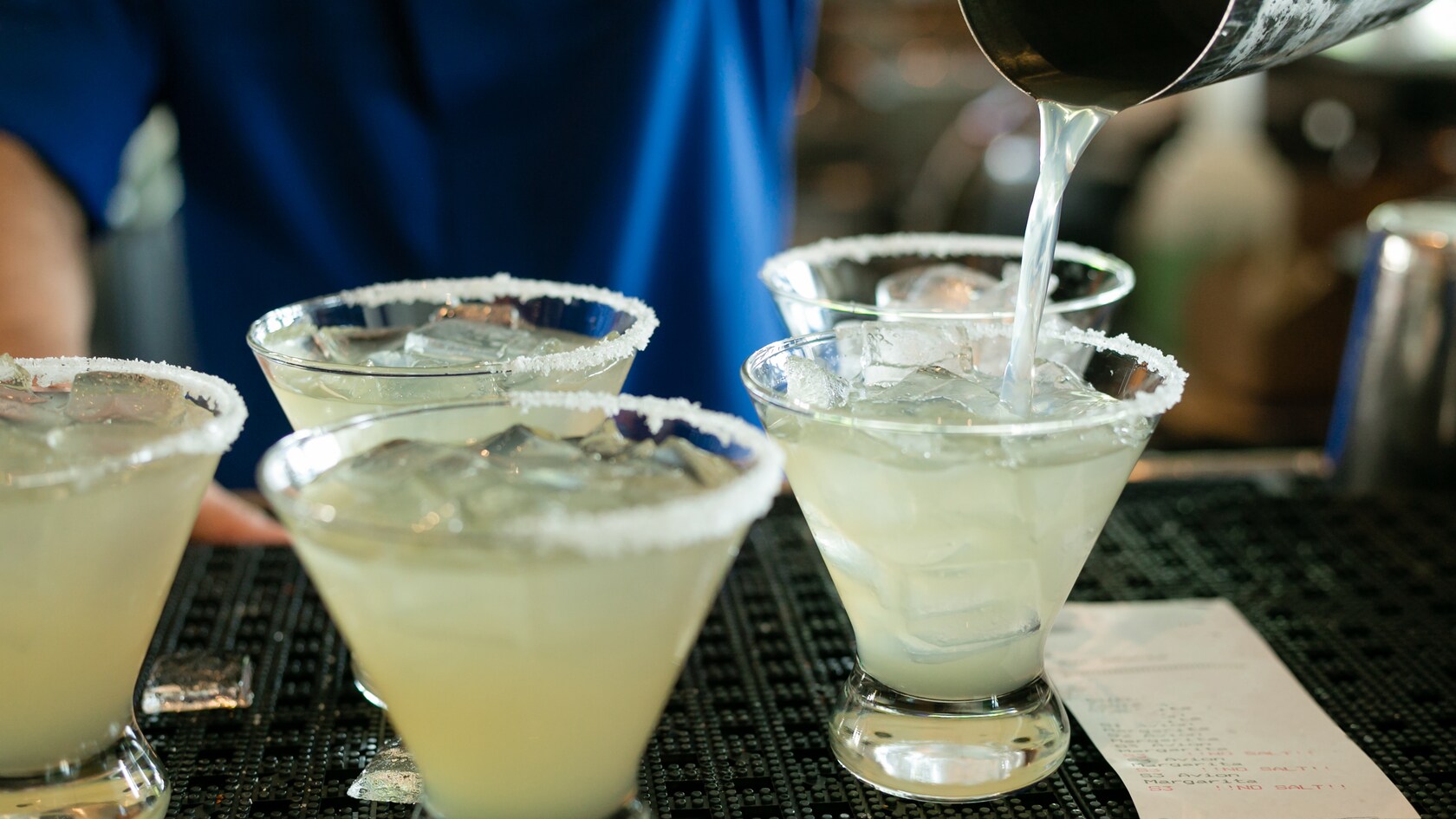 A bartender pouring a freshly made margarita cocktail into glasses with ice and salted rims