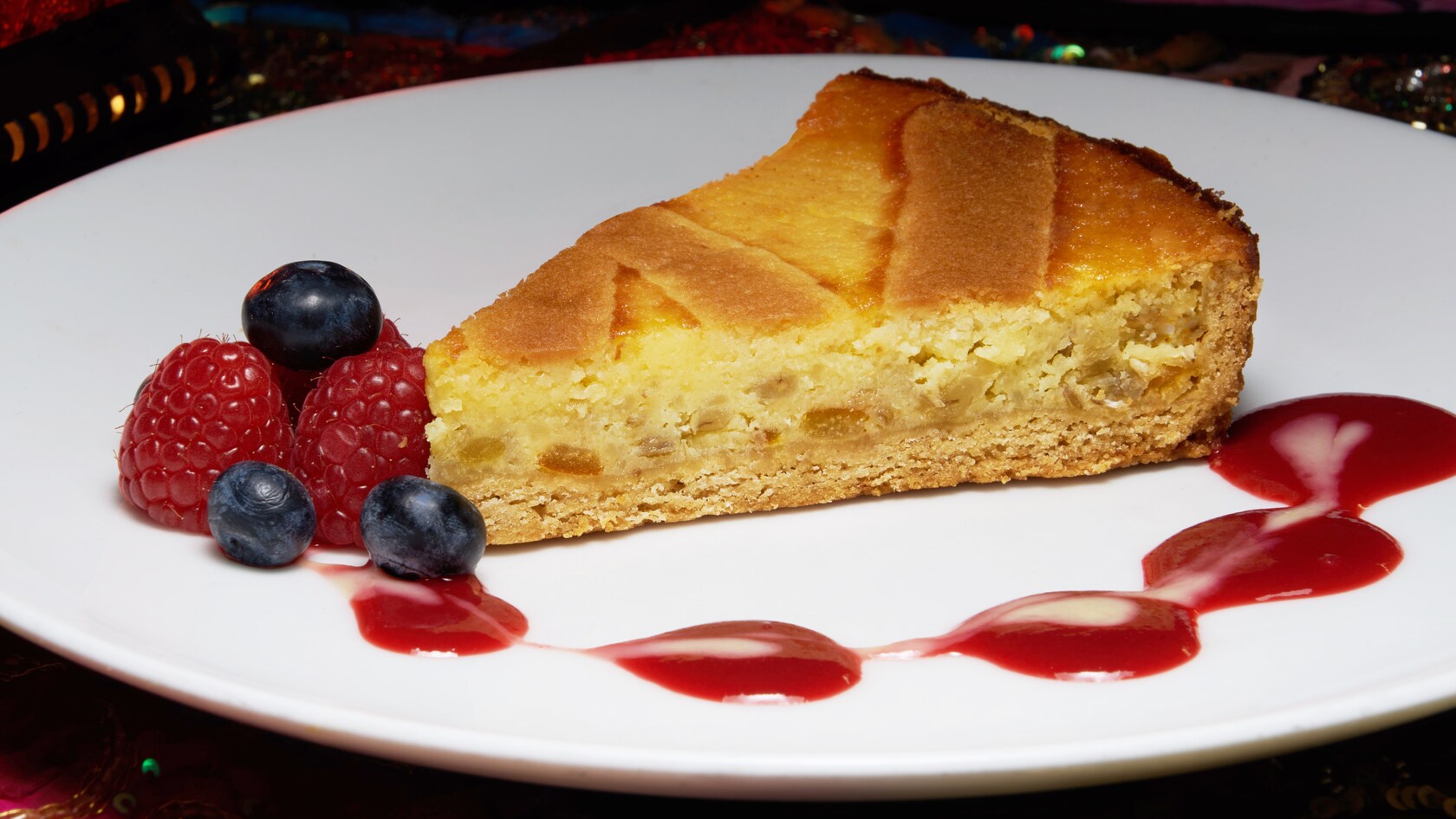 A tart slice with a tender crust on a plate, garnished with berries and a drizzle of fruity sauce