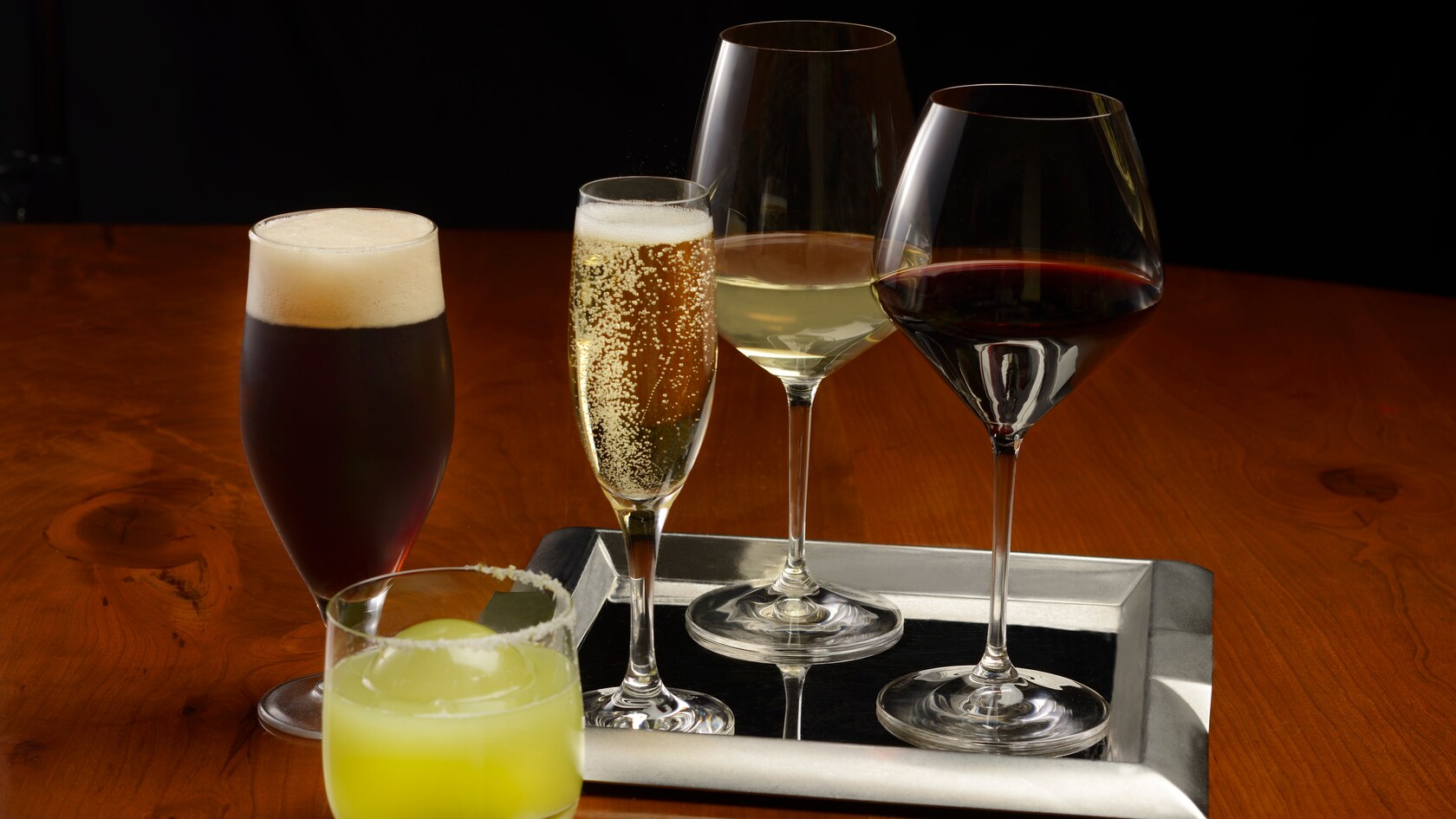 A glass of champagne and 2 glasses of wine on a tray next to a beer and another cocktail