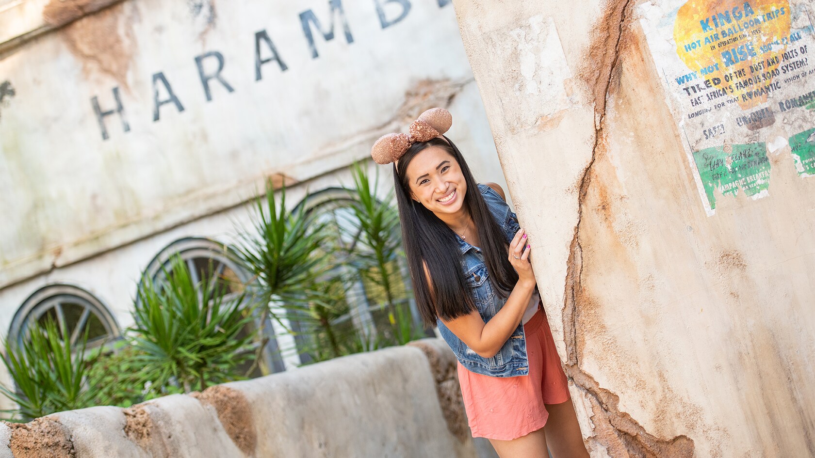 A woman wears a Minnie Mouse ear hat and peaks out from behind a wall, as she stands in front of a weathered building with the word Harambe painted on it