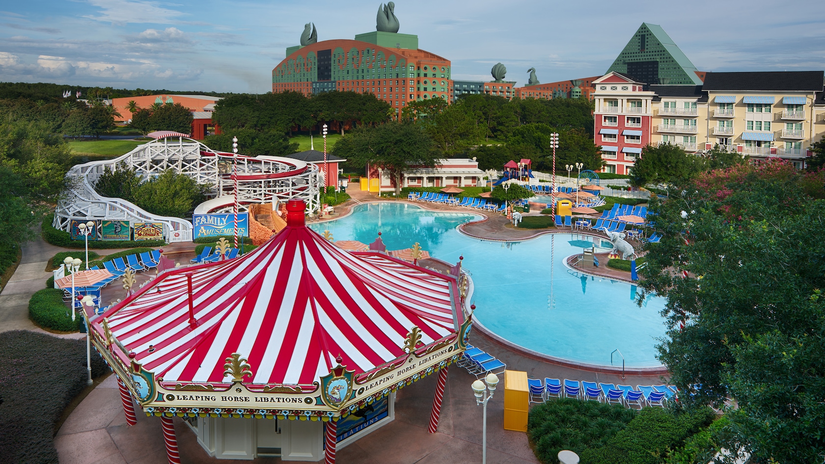 The pool area of Disney's BoardWalk Inn featuring 2 multistory buildings, leafy trees, a carousel shaped snack bar and a waterslide fashioned to look like a roller coaster