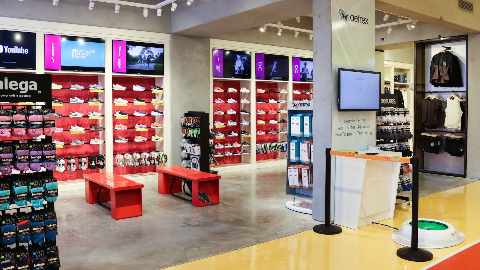 El interior de Fit2Run con zapatos deportivos exhibidos en la pared, estantes con accesorios para correr, bancos para sentarse, monitores de TV y un área dedicada al dispositivo de escaneo de pies Aetrex