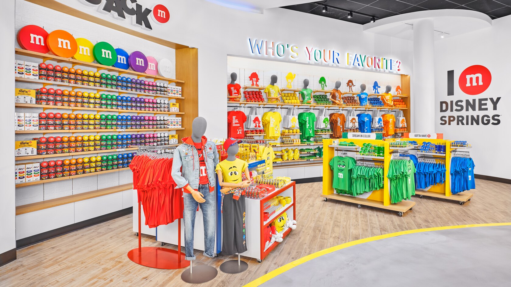 Un magasin débordant de produits M and M, dont des chandails, des vestes, des tasses et des emballages de bonbons. Différentes enseignes indiquant Who’s Your Favorite, Dream in Color, Gotta Have M, et M Disney Springs.