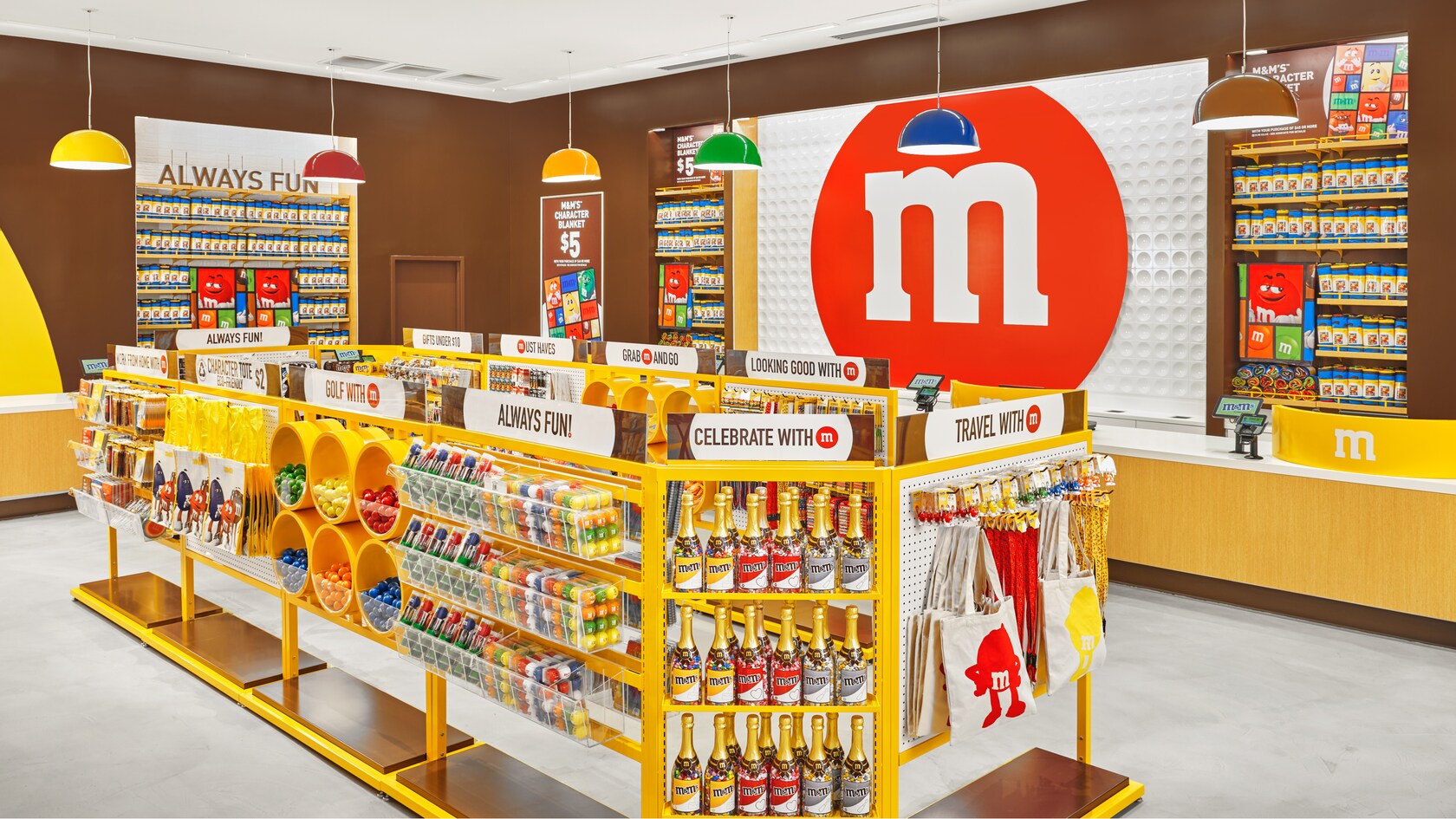 Une caisse et une file bordée de présentoirs avec des produits M and M, dont des sacs fourre-tout, des balles de golf et des bouteilles de champagne remplies de bonbons. Différentes enseignes indiquant Work from Home with M, Character Tote Eco Friendly, Gold with M, Always Fun, Celebrate with M, Travel with M, Looking Good with M, Grab M and Go, Must Haves, Gifts Under $10, et M and Ms Character Blanket $5.