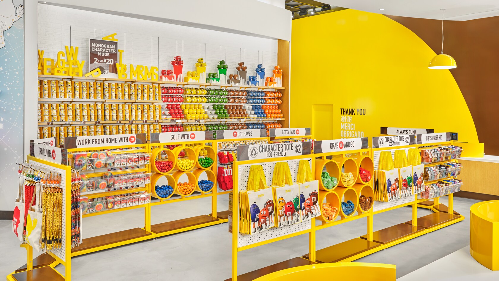 La file d’une caisse bordée de présentoirs avec des produits M and M, dont des sacs fourre-tout, des balles de golf, des tasses et des chaussettes. Différentes enseignes indiquant Monogram Character Mugs 2 for $20, Work from Home with M, Character Tote Eco Friendly, Gold with M, Always Fun, Travel with M, Looking Good with M, Grab M and Go, Must Haves, Gifts Under $10 et Thank You, Merci, Obrigado