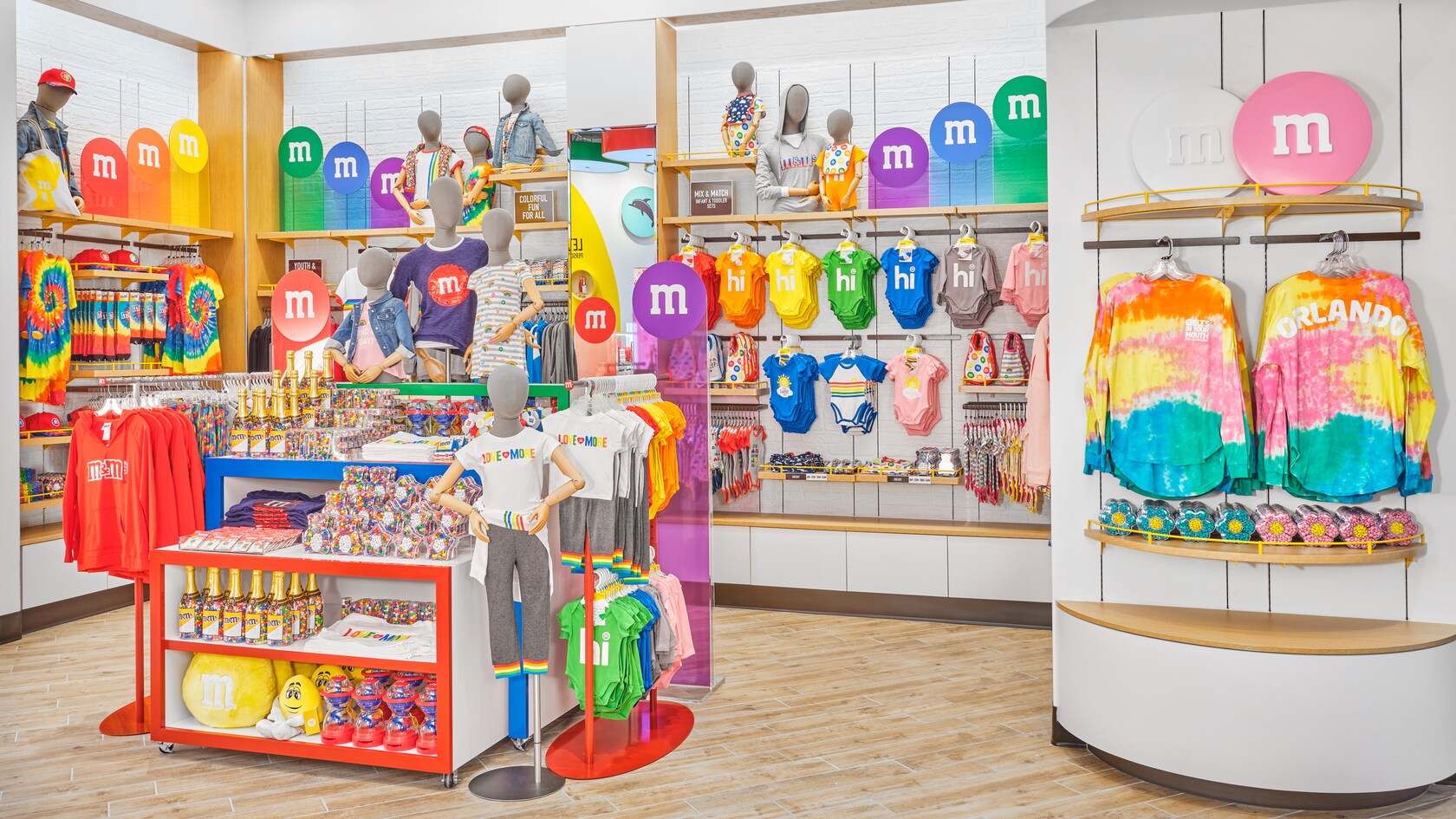 Des présentoirs avec des produits M and M pour les enfants, comme des chandails, des pantalons, des bavettes, des grenouillères, des chapeaux, des jouets, des coussins et différents contenants remplis de bonbons, dont des bouteilles de champagne