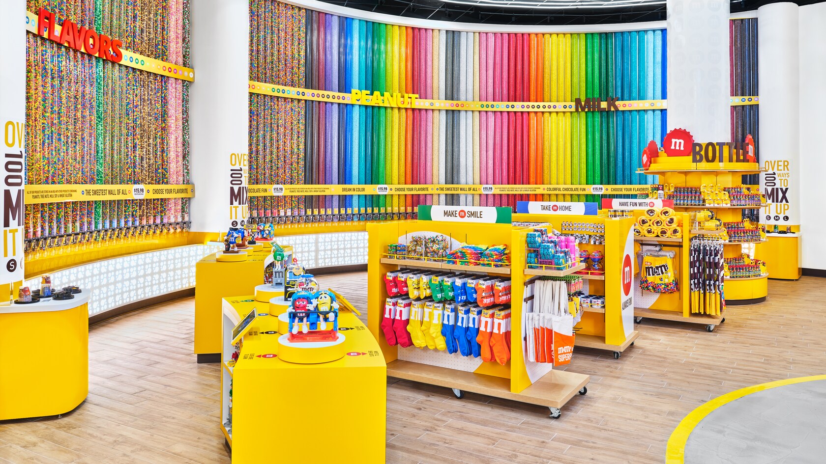 De grands contenants cylindriques remplis de bonbons recouvrent le mur du fond dans un magasin débordant de produits M and M, dont des jouets, des chaussettes, des sacs fourre-tout et des serviettes. Des enseignes indiquant Flavors, Peanut, Milk, Make M Smile, Take M Home, Have Fun with M, et Bottle.