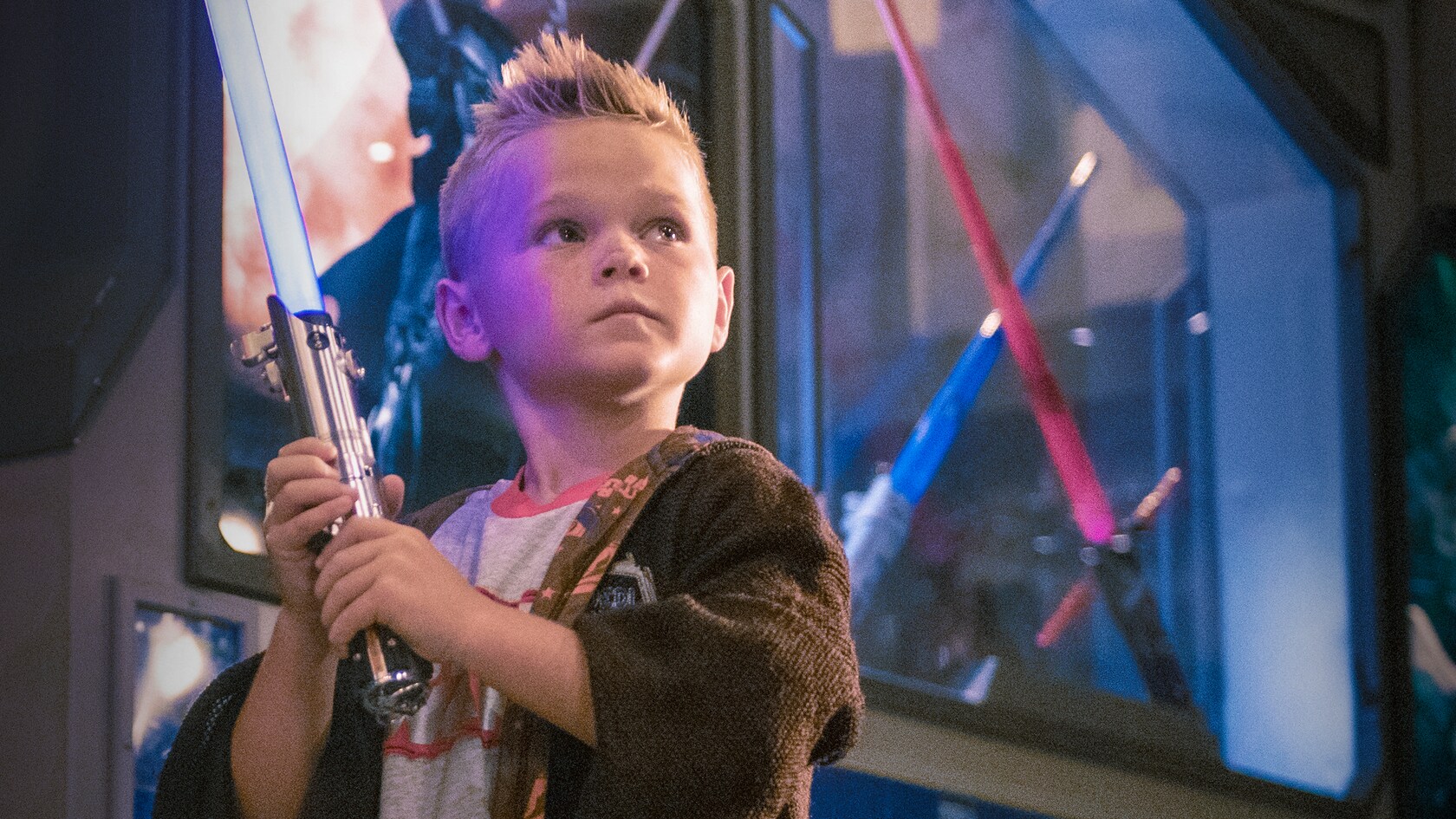 Amidst store displays, a young boy in a Jedi robe poses with his lightsaber while looking off into the distance