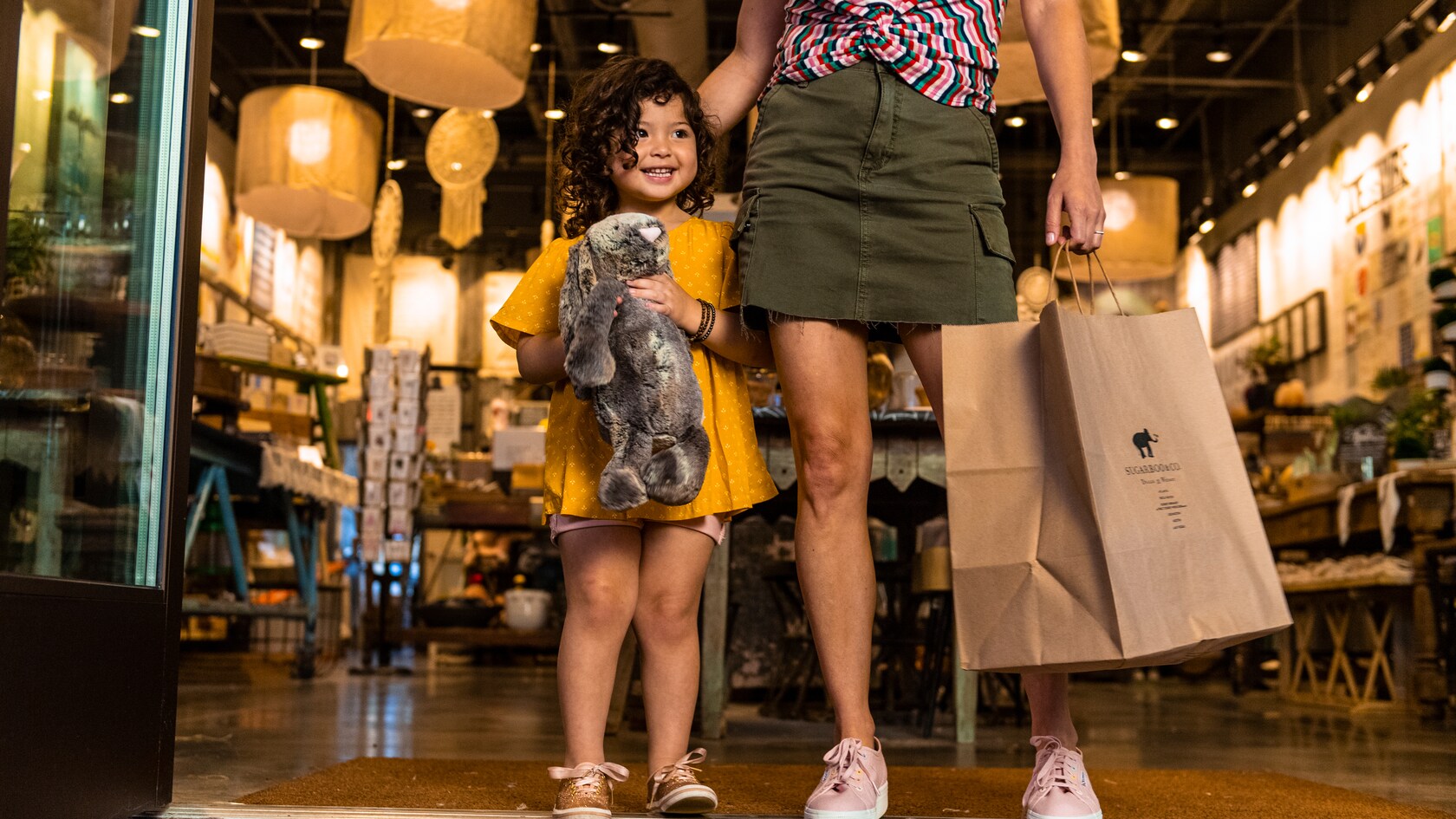 Uma menina segurando um coelho de pelúcia e caminhando por uma loja com a mãe