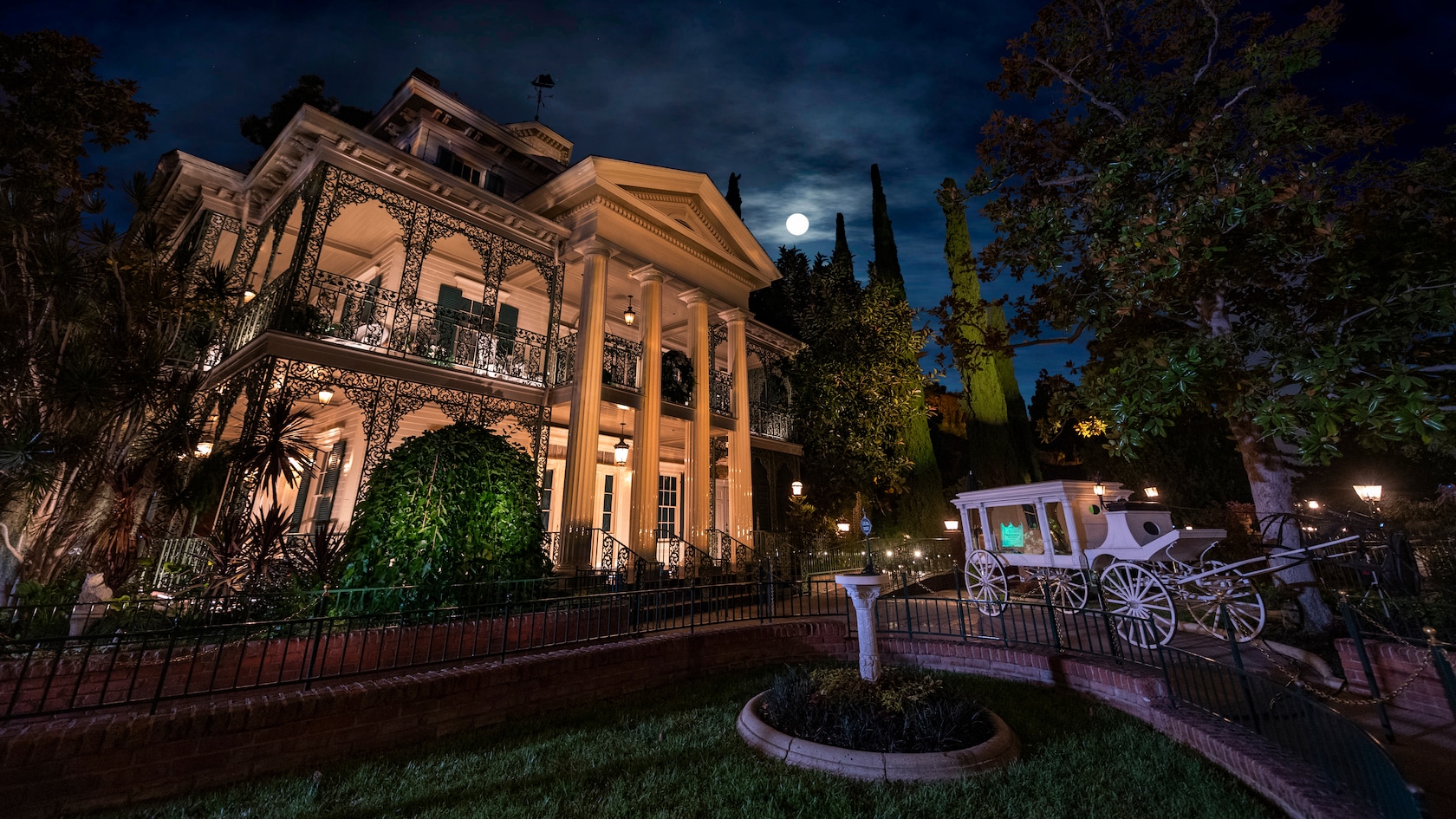 Under moonlit skies, a horse drawn carriage sits outside the eerily illuminated exterior of the Haunted Mansion 
