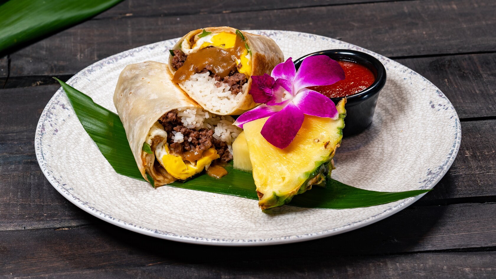 Un burrito relleno de arroz, carne molida, huevo frito y salsa gravy en un plato con una taza de salsa, rebanadas de piña y una flor de orquídea