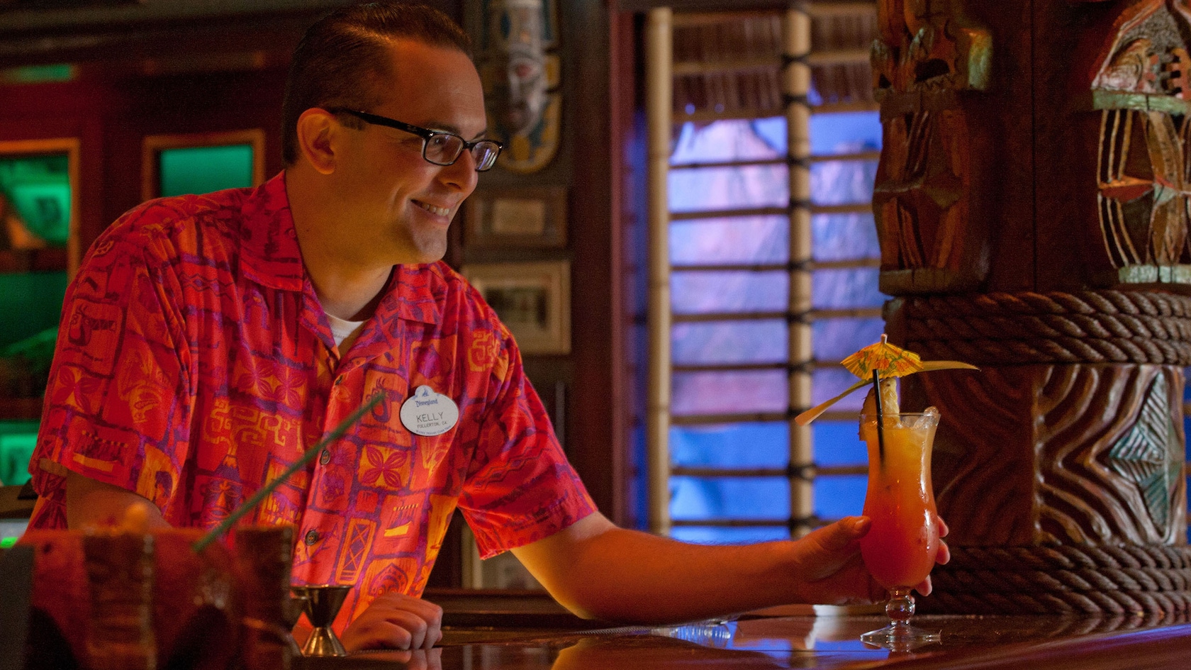 A bartender serves a tropical cocktail topped with a pineapple slice and mini umbrella to a Guest.