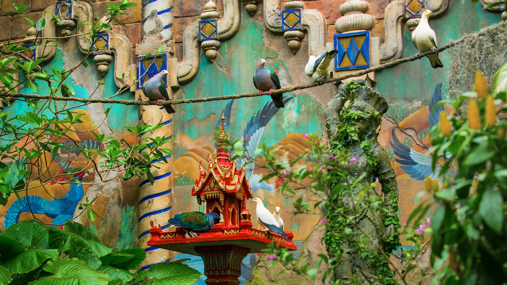 Exotic birds eat from a ornate birdhouse next to the ruins of a colorful wall