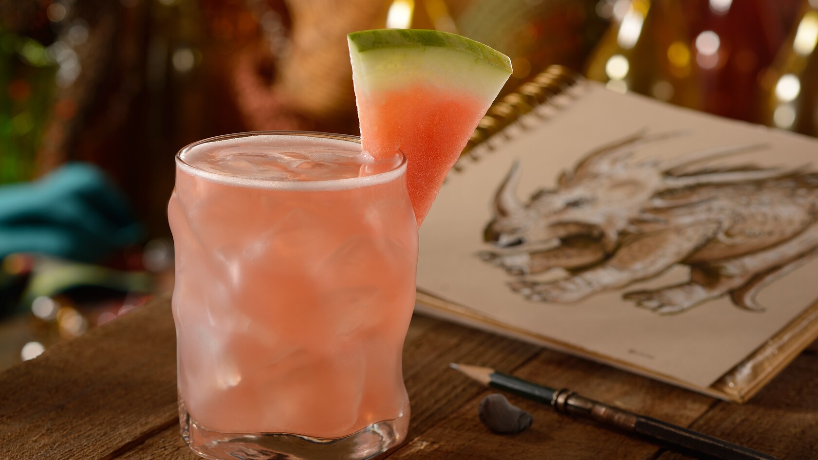An icy glass with a Hightower Rita cocktail on a table beside a pencil and a sketchbook open to a drawing of a triceratops