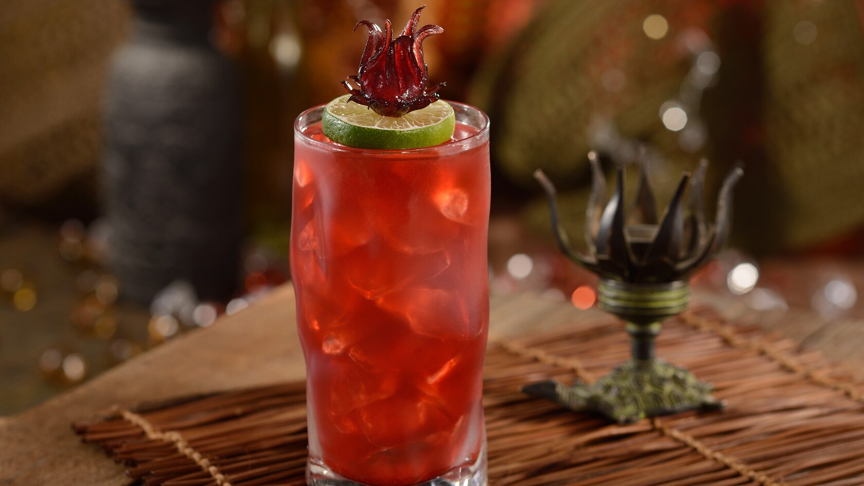 A tall icy glass with a Jenn’s Tattoo cocktail atop a woven reed mat beside an ornate Polynesia styled candle holder