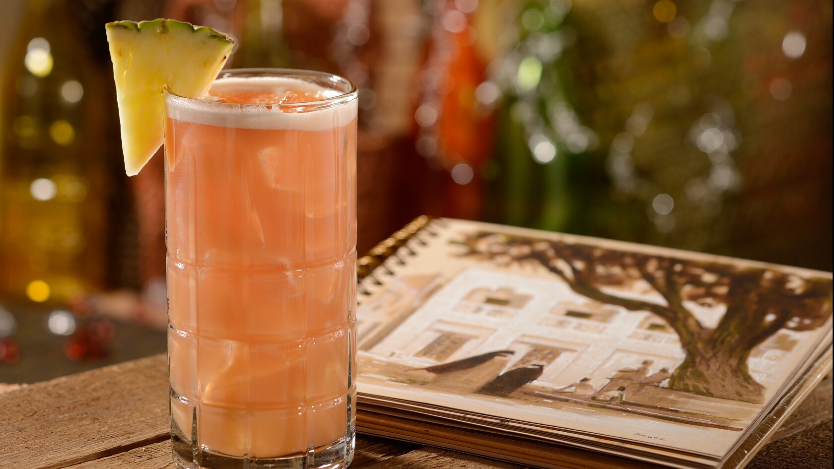 A tall icy glass with a Lamu Libation cocktail garnished with a pineapple wedge on a table beside a sketchbook open to a page with a drawing of an African village square