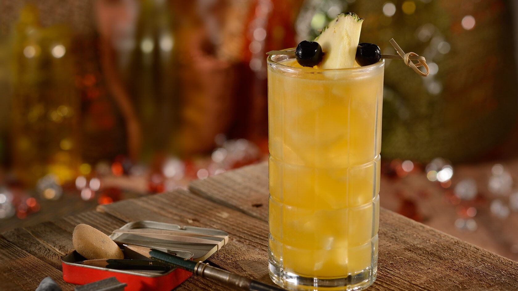 A tall icy glass with a Leaping Lizard cocktail garnished with a pineapple wedge skewered between two blueberries on a table beside a tin box and drawing implements