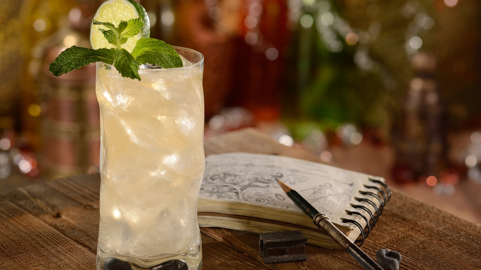 A tall icy glass with a Snow Leopard Salvation cocktail garnished with mint leaves and a lime slice on a table beside a sketch book open to an ornate drawing of a mythological scene