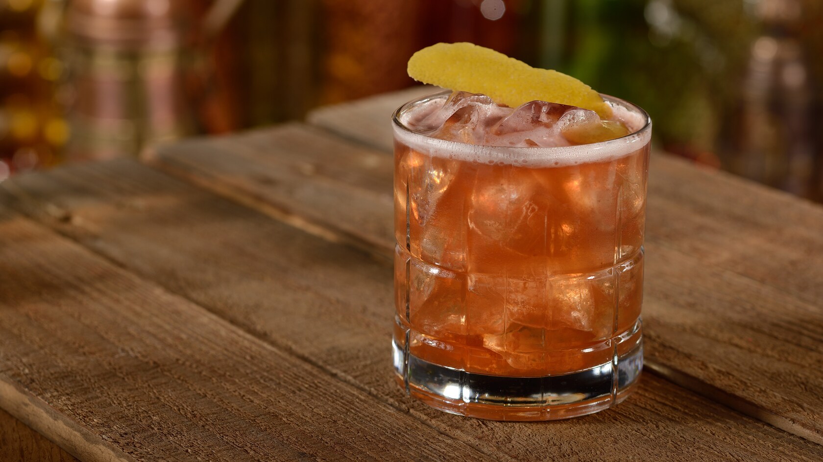 An icy glass with a Tempting Tigress cocktail garnished with a slice of  lemon peel on a wooden table