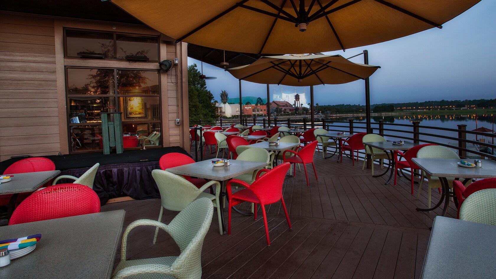 On the lakeside deck, large umbrellas hang above tables with red and white wicker chairs 