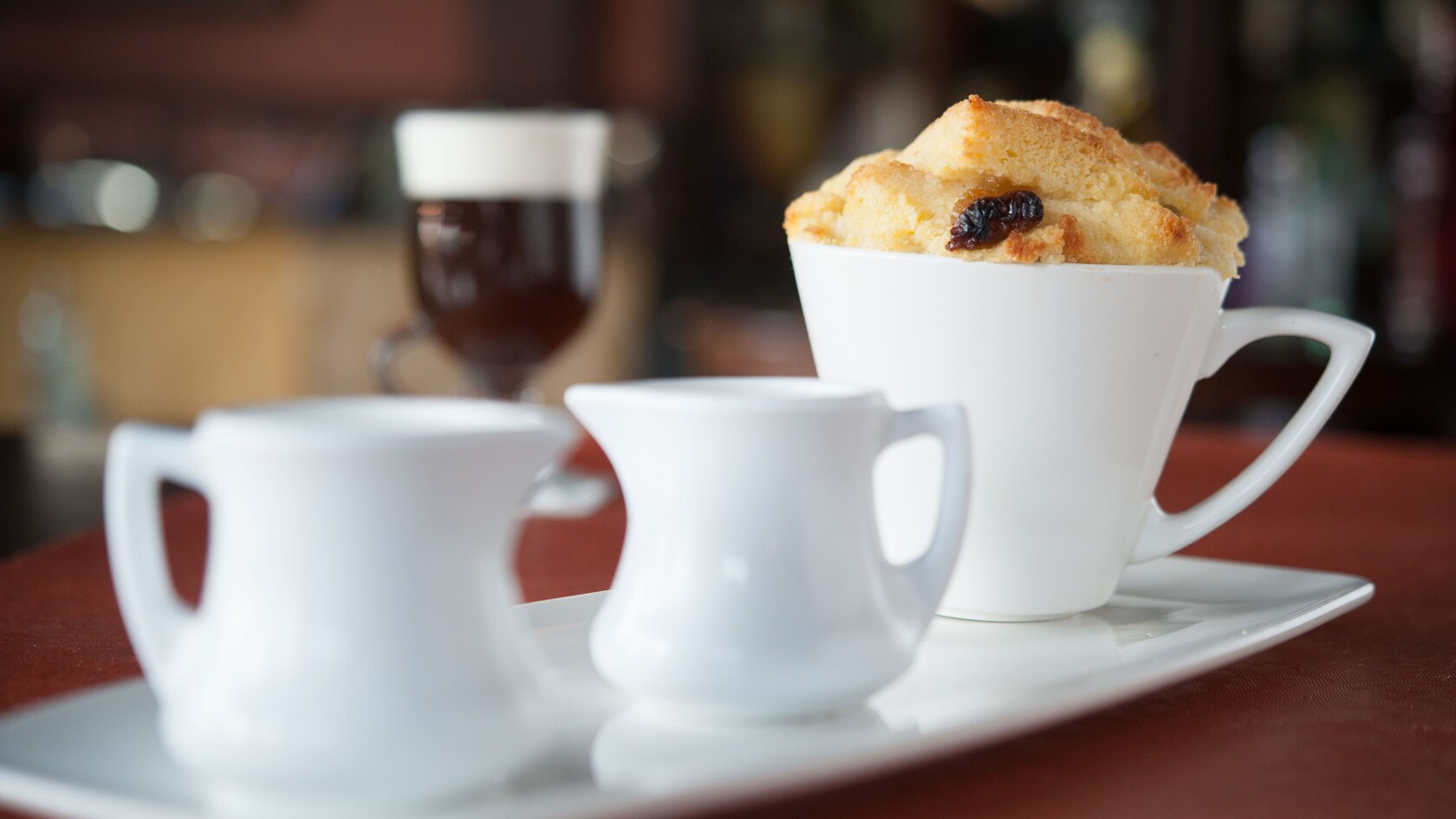 Bread and butter pudding in a ramekin next to 2 small jugs of sauce