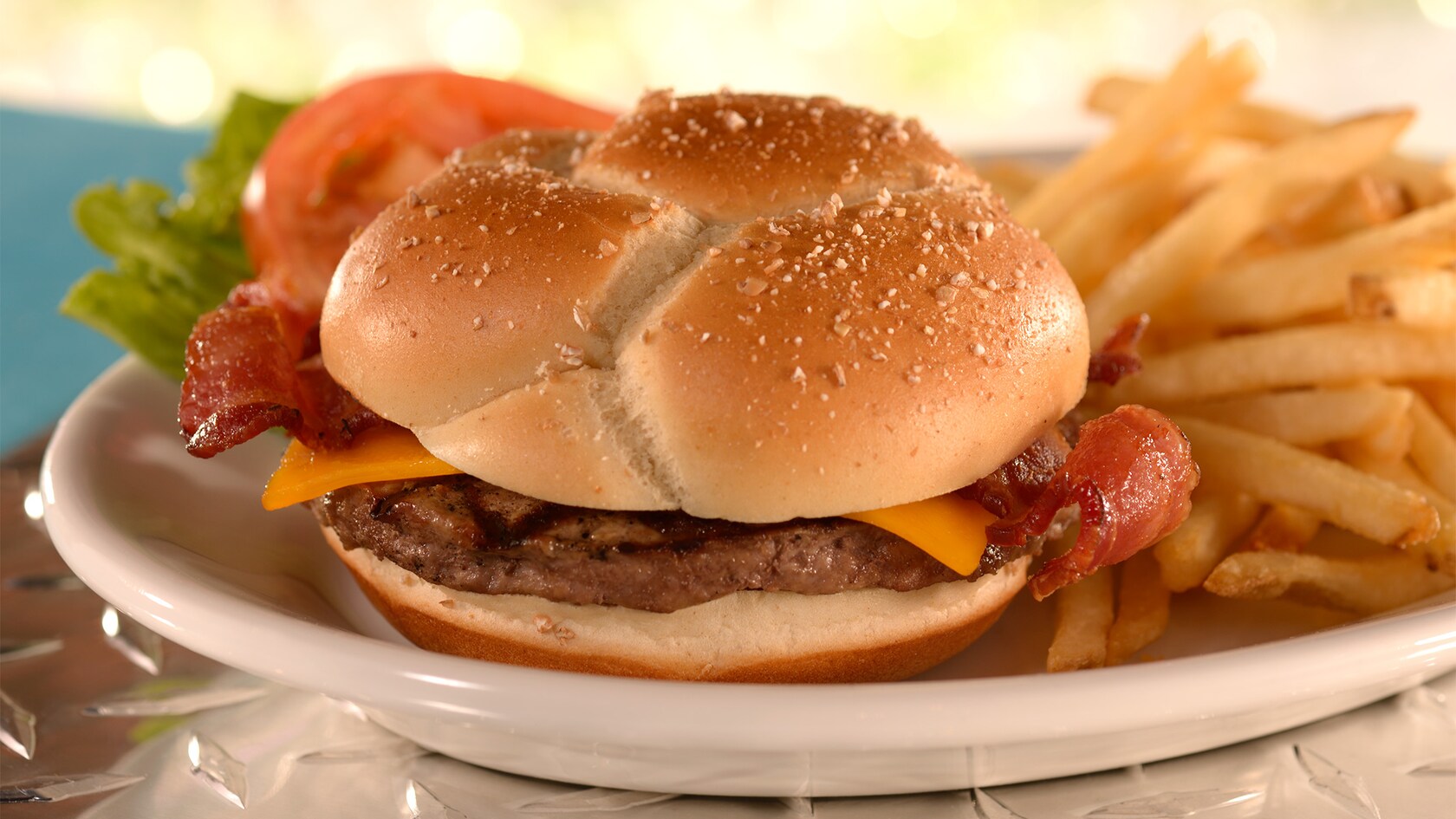 A bacon cheese burger on a bun served with a lettuce leaf, a tomato slice and an order of French fries