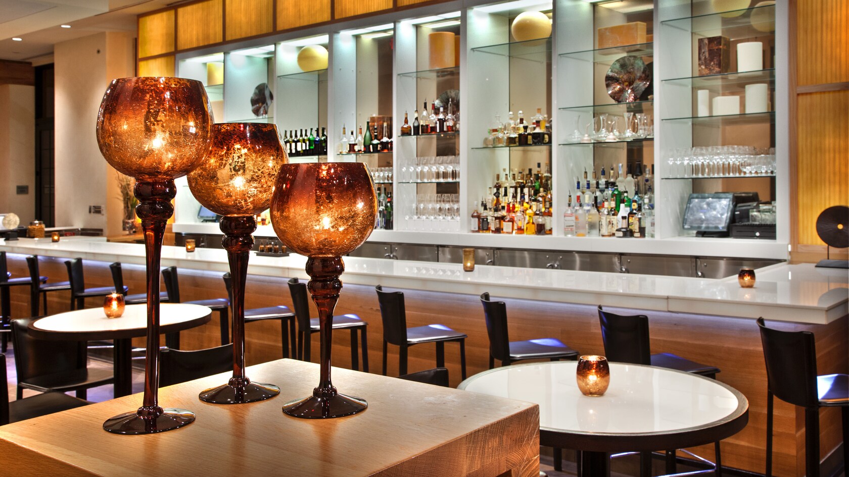 Tres portavelas de vidrio color ámbar en primer plano, con bar, mesas y taburetes en el fondo