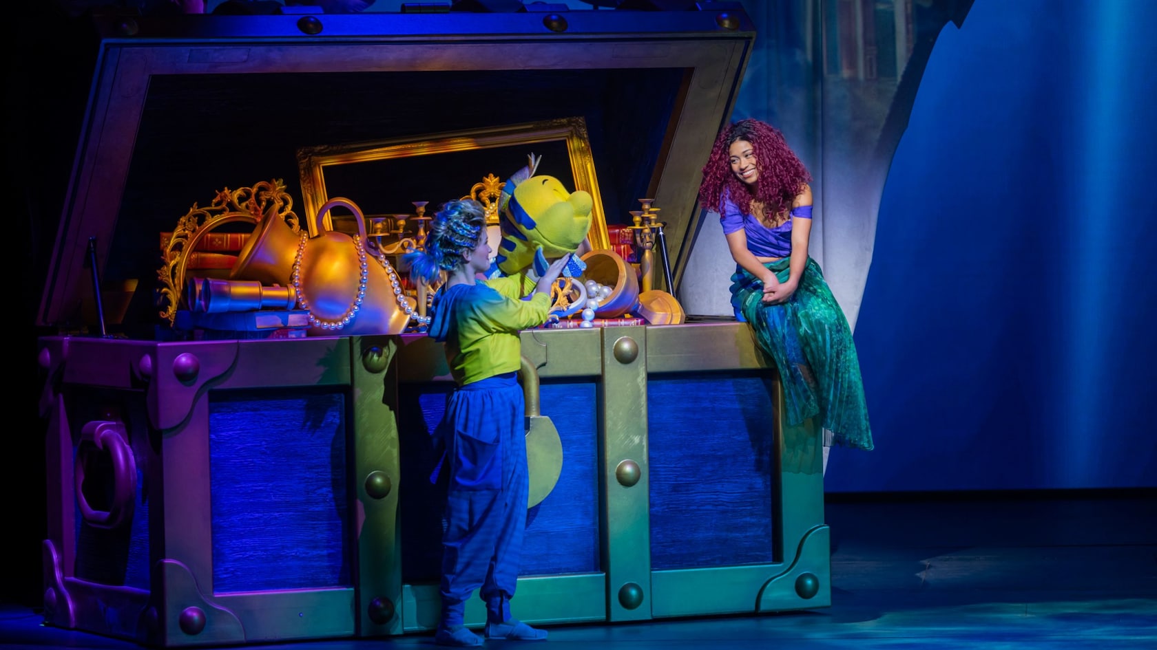 A stage performance of The Little Mermaid reveals Ariel sitting on a giant treasure chest