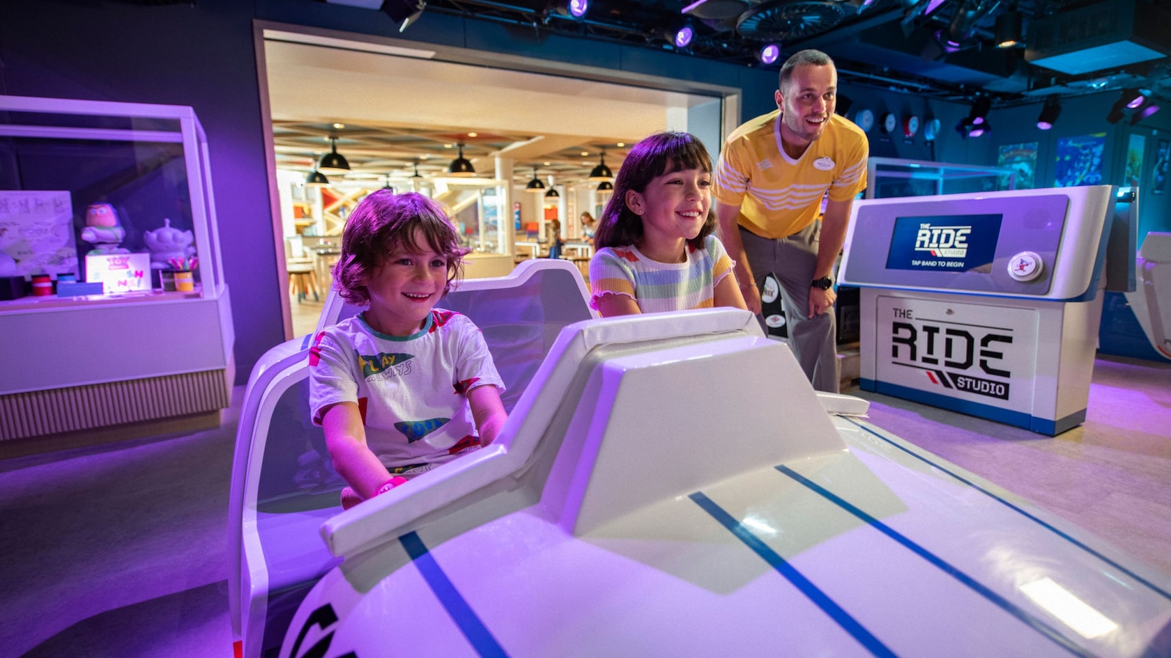 2 children sitting in a space ship simulator while a Disney youth counselor looks on