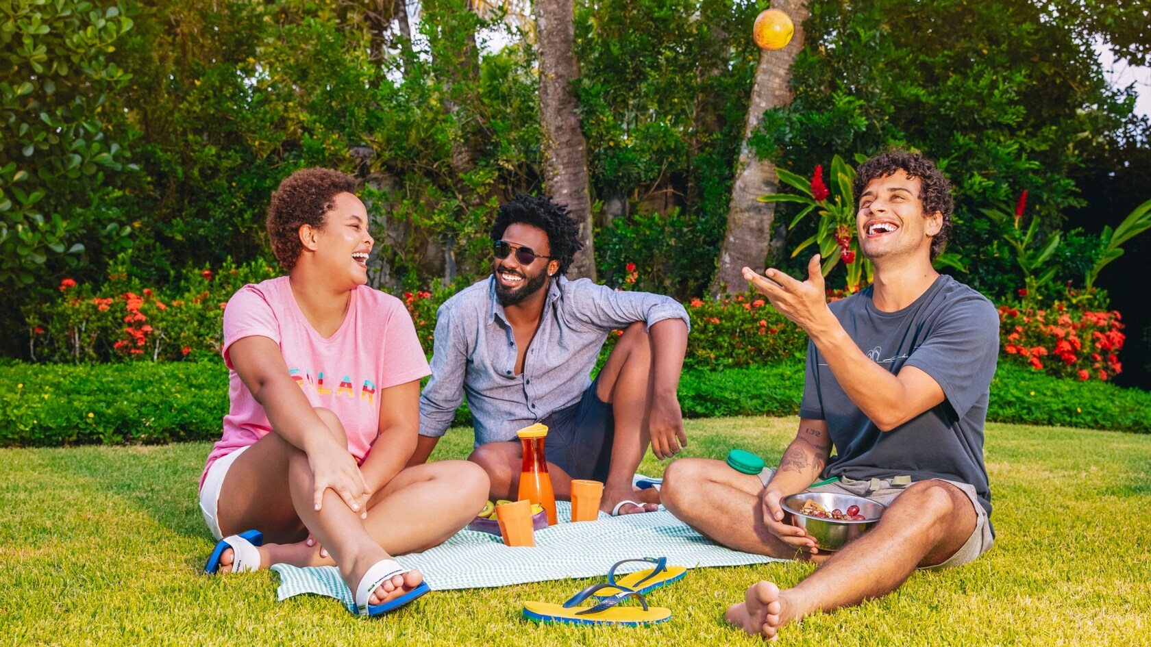 Three friends sit outside having a picnic while one throws an apple in the air