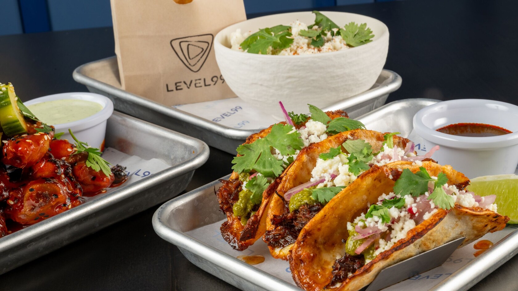 Tacos from Level99 Disney Springs topped with cheese, guacamole and cilantro served on a tray alongside glazed skewers and a grain bowl