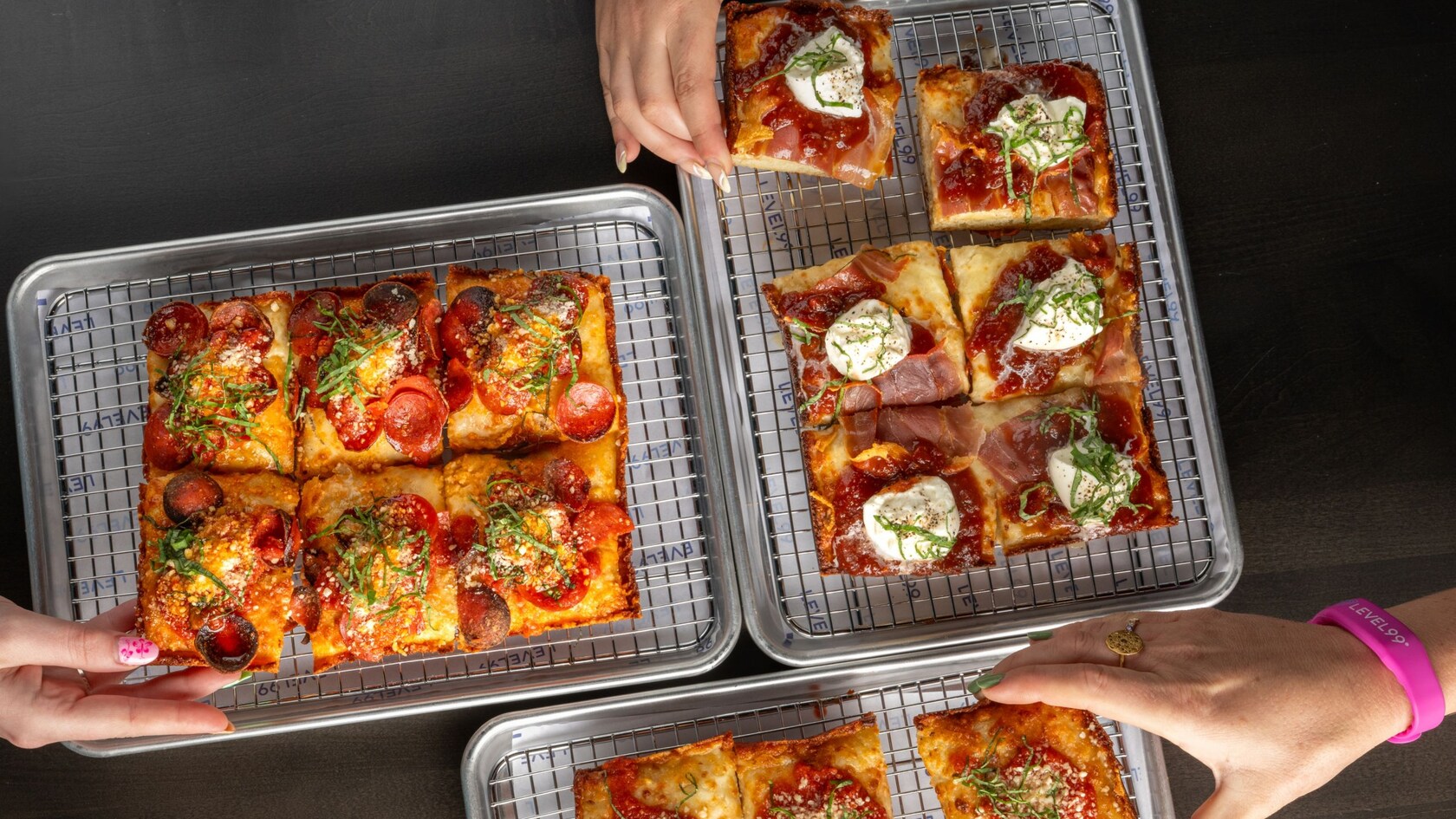 Hands reaching for 3 trays of square cut pizza with varied toppings