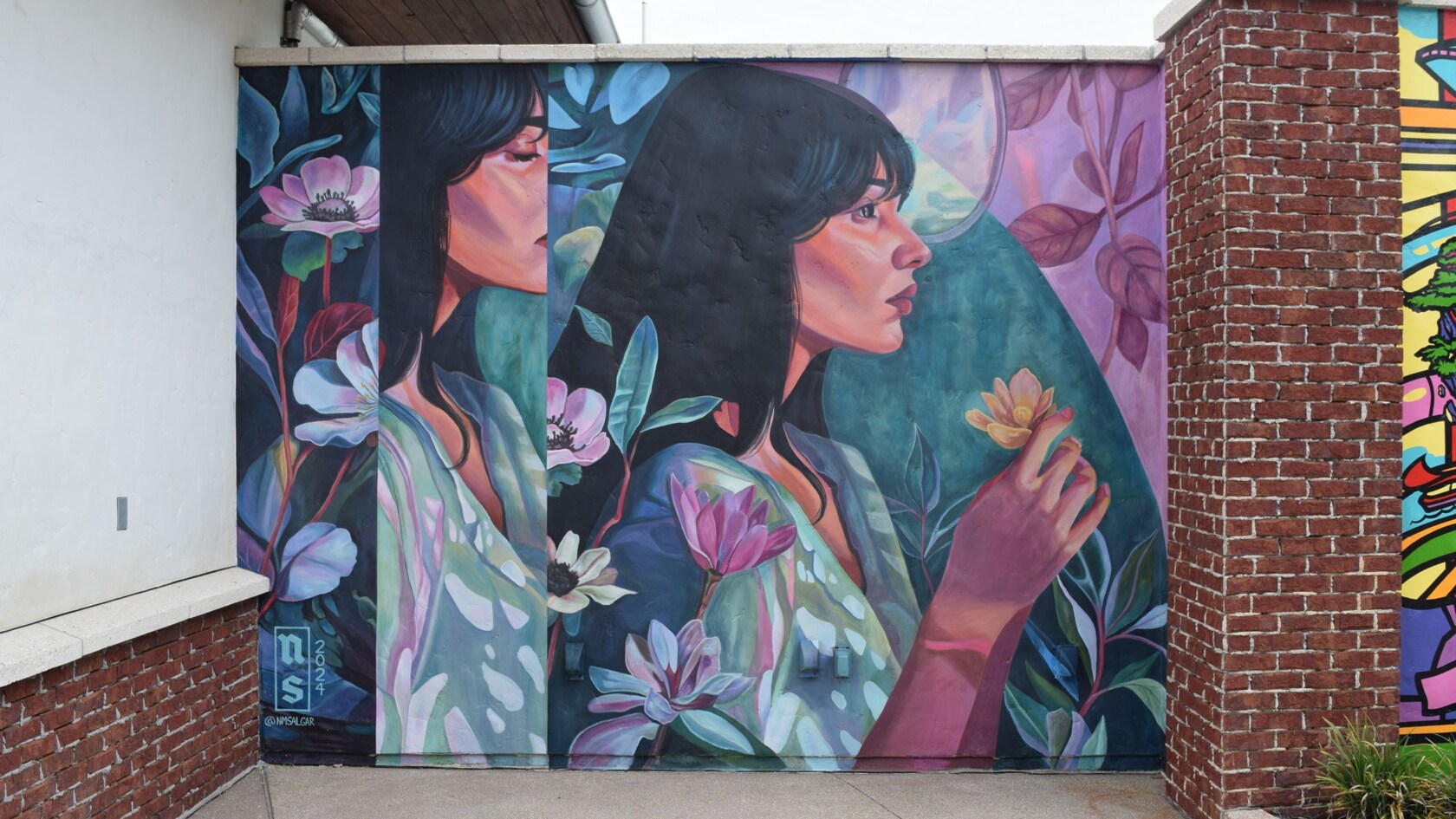 Un mural sobre una pared de ladrillo de Nicole Salgar con dos perfiles de una mujer rodeada de flores y follaje, sosteniendo una flor