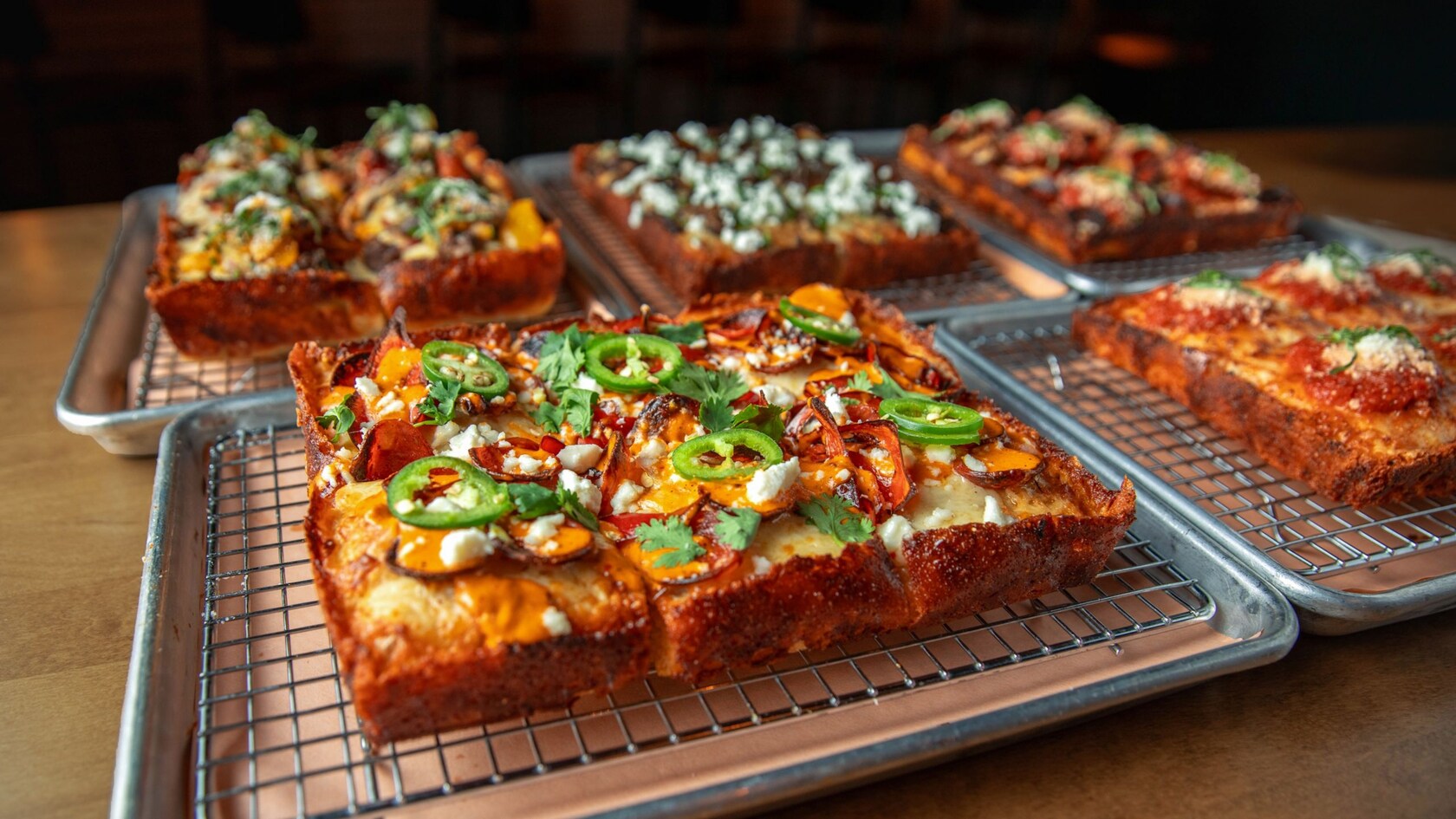 Several pans of Detroit style pizza, featuring one with jalapenos, cilantro, pepperoni and crumbled cheese