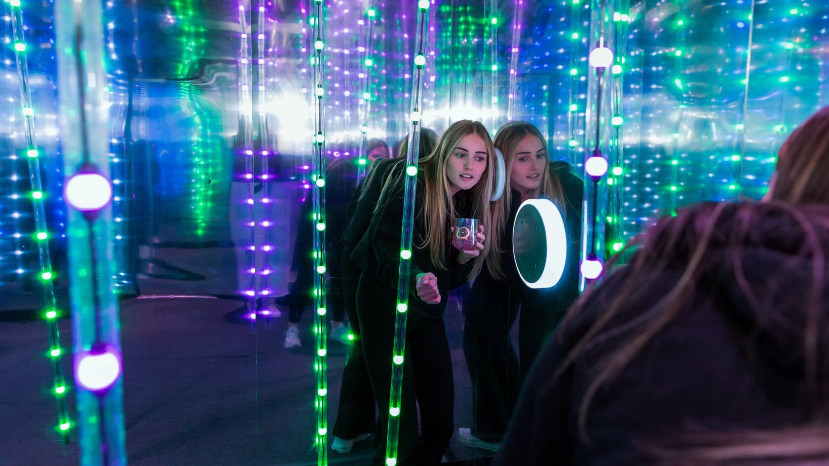 A Guest holding a beverage and listening to a speaker in a mirrored room with strings of lights