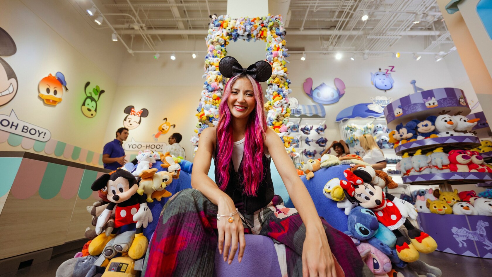 A woman wearing a Minnie Mouse ear headband sitting on a throne decorated with Disney Character plush toys inside the Disney Ever After store
