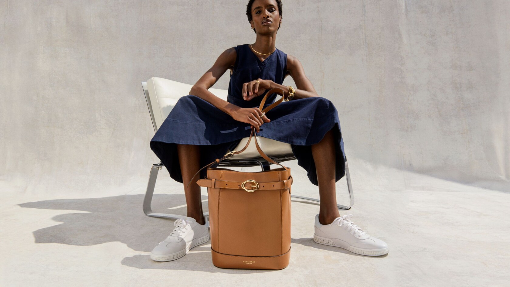 A woman seated in a chair wearing Cole Haan sneakers with a leather bucket bag at her feet
