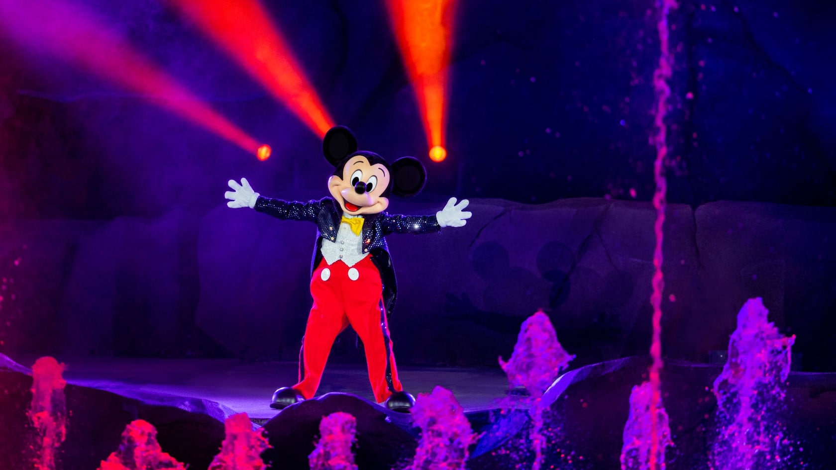 Mickey Mouse stands on a stage near water jets
