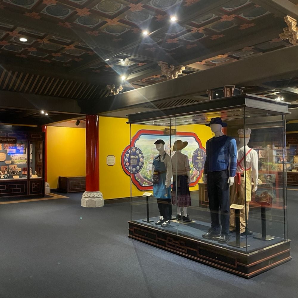 The interior of House of Whispering Willows at Shanghai Disney Resort featuring display cases with Disney artwork and 4 mannequins in vintage costumes