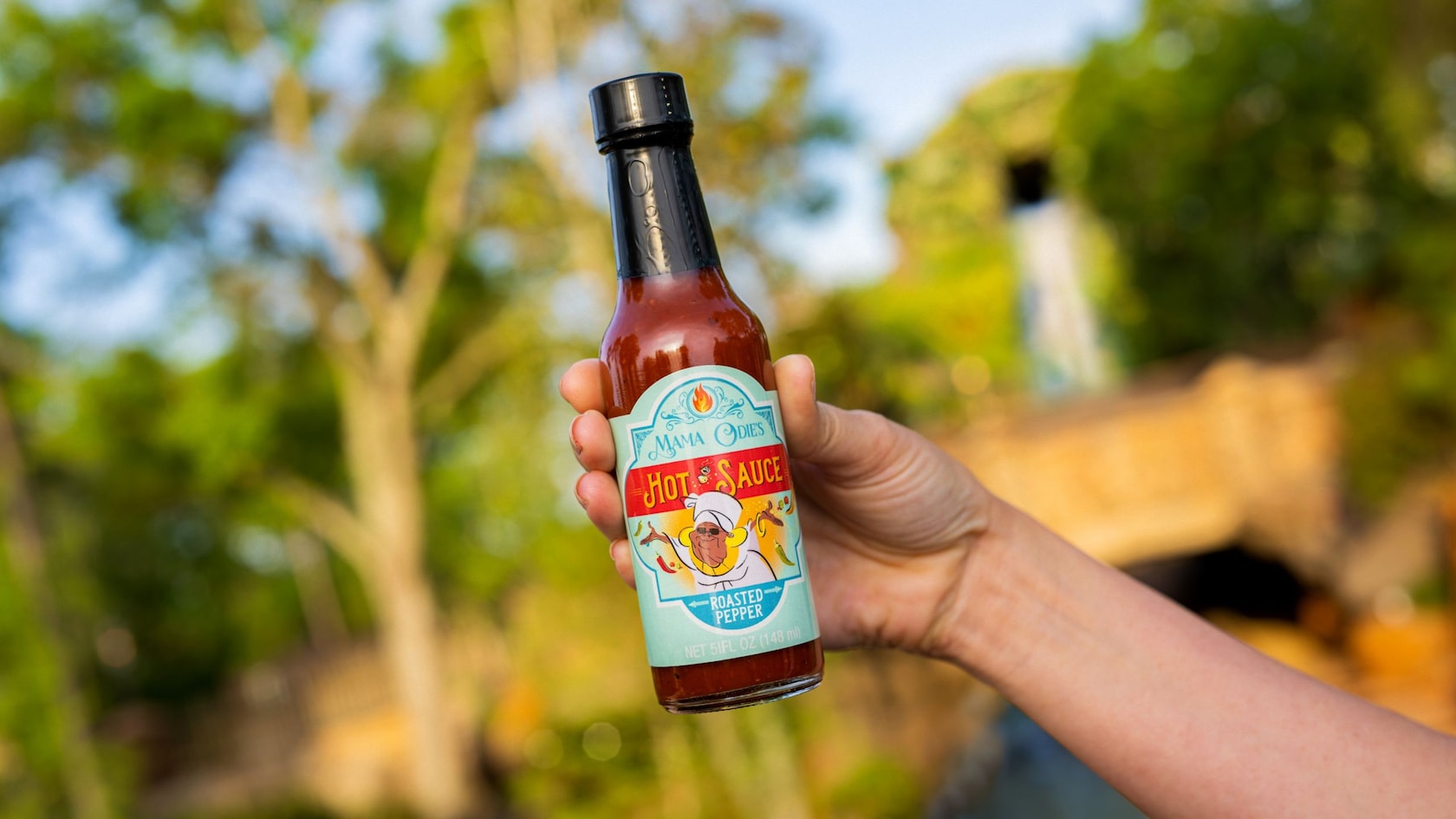 A hand holding a bottle of hot sauce with a label that says, ‘Mama Odies Hot Sauce Roasted Pepper’