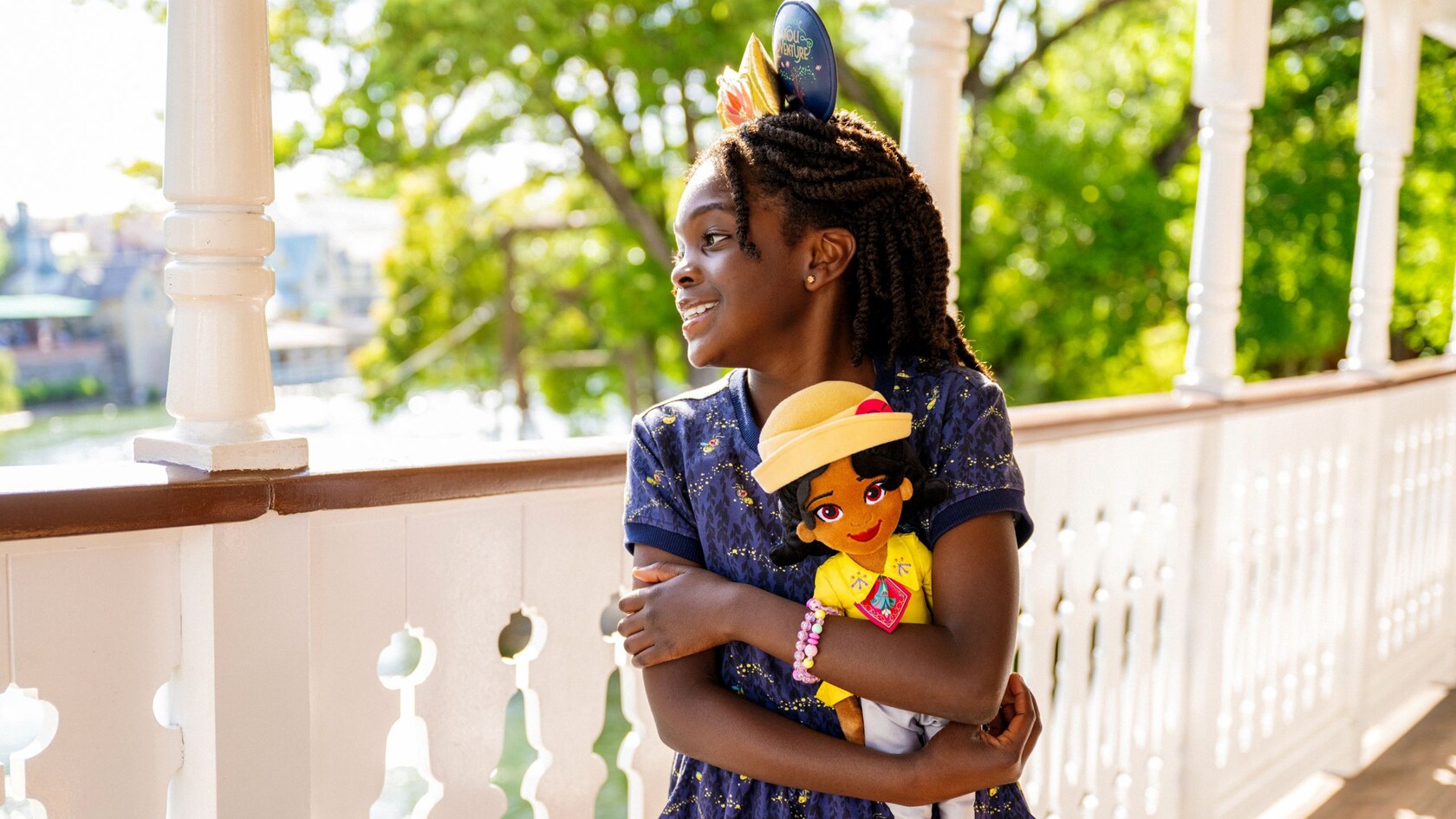 A girl hugging a Tiana plush doll while wearing a Tiana’s Bayou Adventure Ear Headband and a dress featuring Ray from ′The Princess and the Frog′