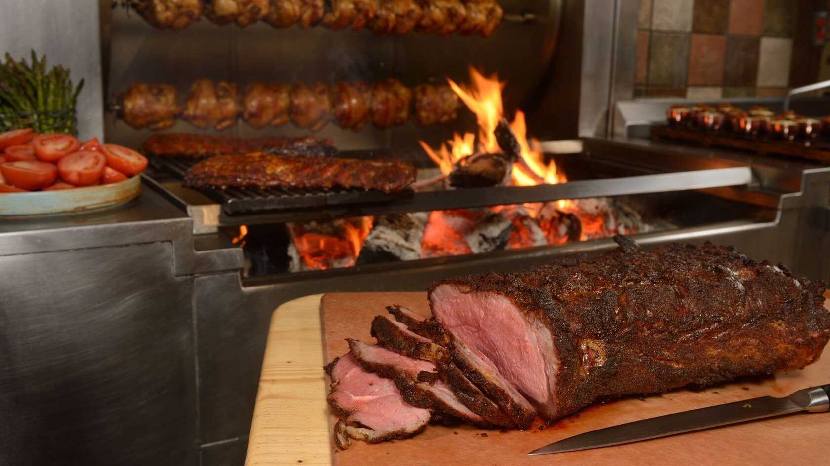 Several chickens cooking on a rotisserie, slabs of meat on a grill and a large roast beef resting on a cutting board