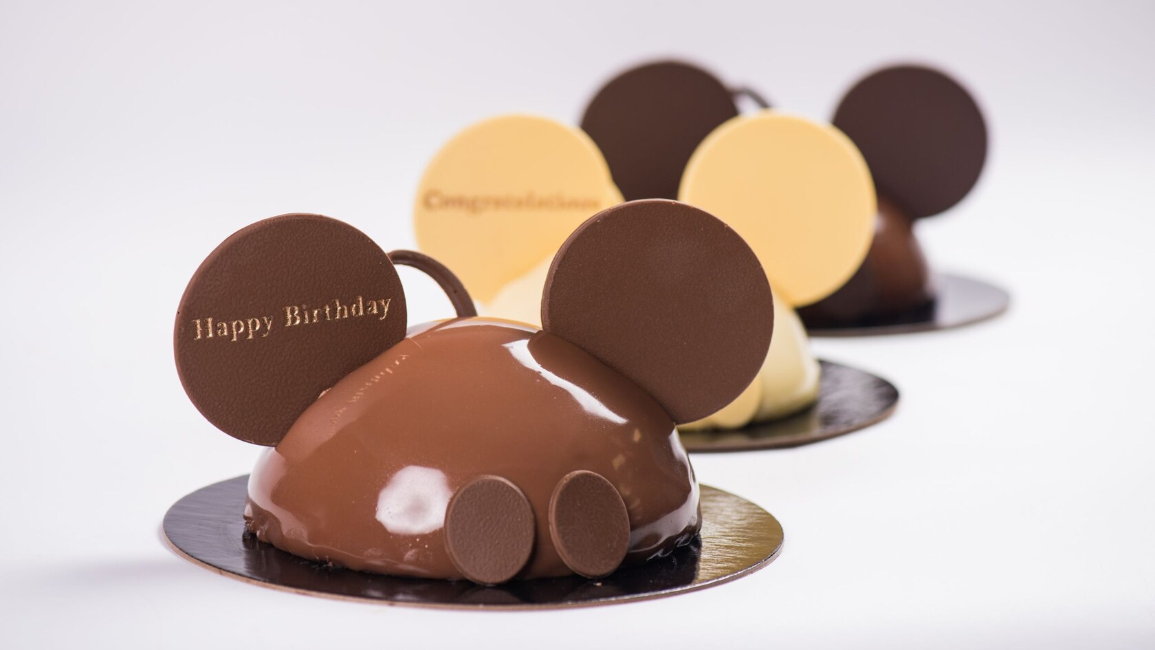 Tres pasteles de celebración de Mickey, de chocolate con leche, chocolate blanco y chocolate amargo, con forma de sombrero con orejas de Mickey Mouse, con orejas, botones y el texto ‘Happy Birthday’ en la oreja del pastel delantero