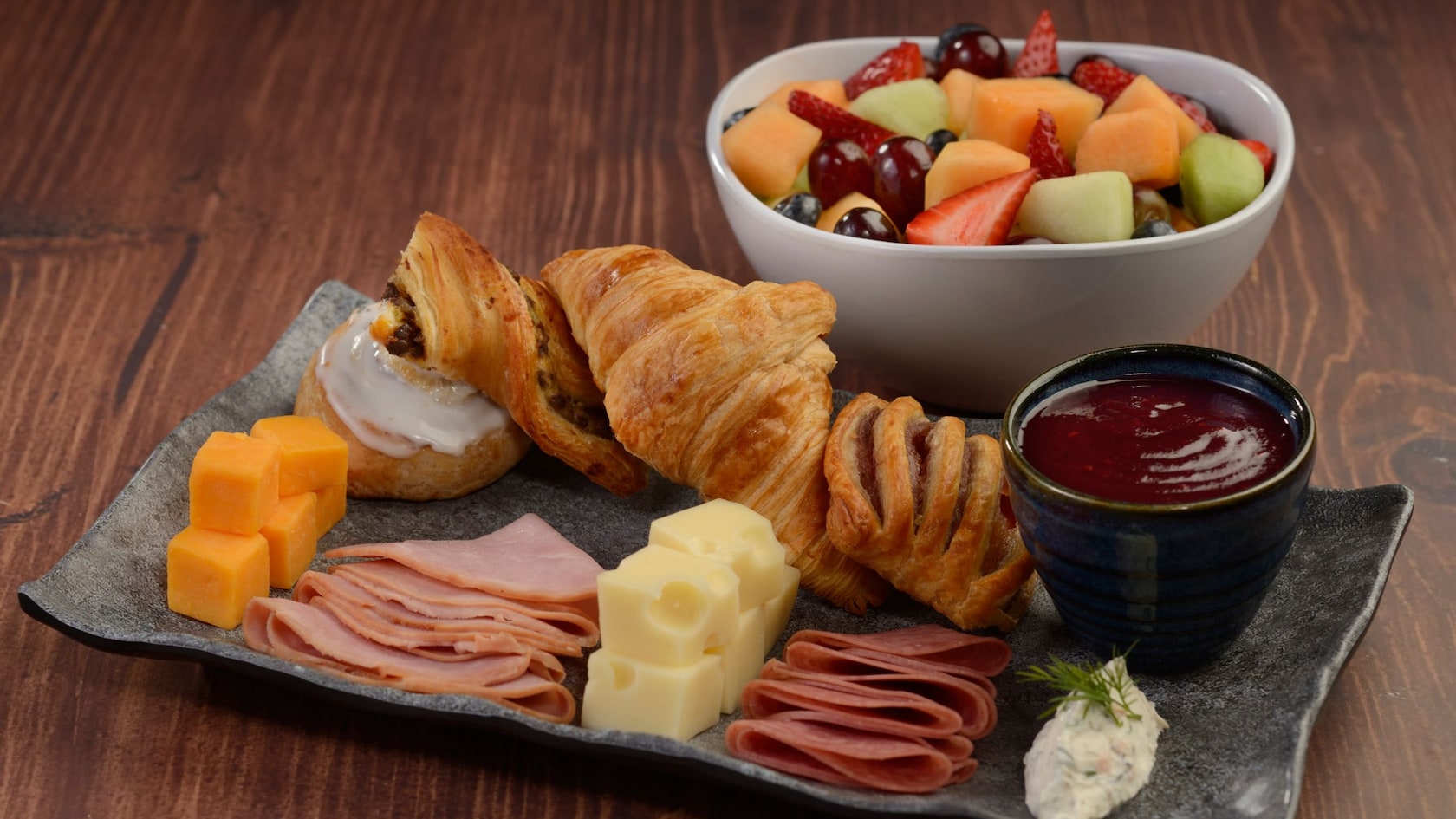 A bowl of mixed fruit and a platter of pastries, cheeses and meats