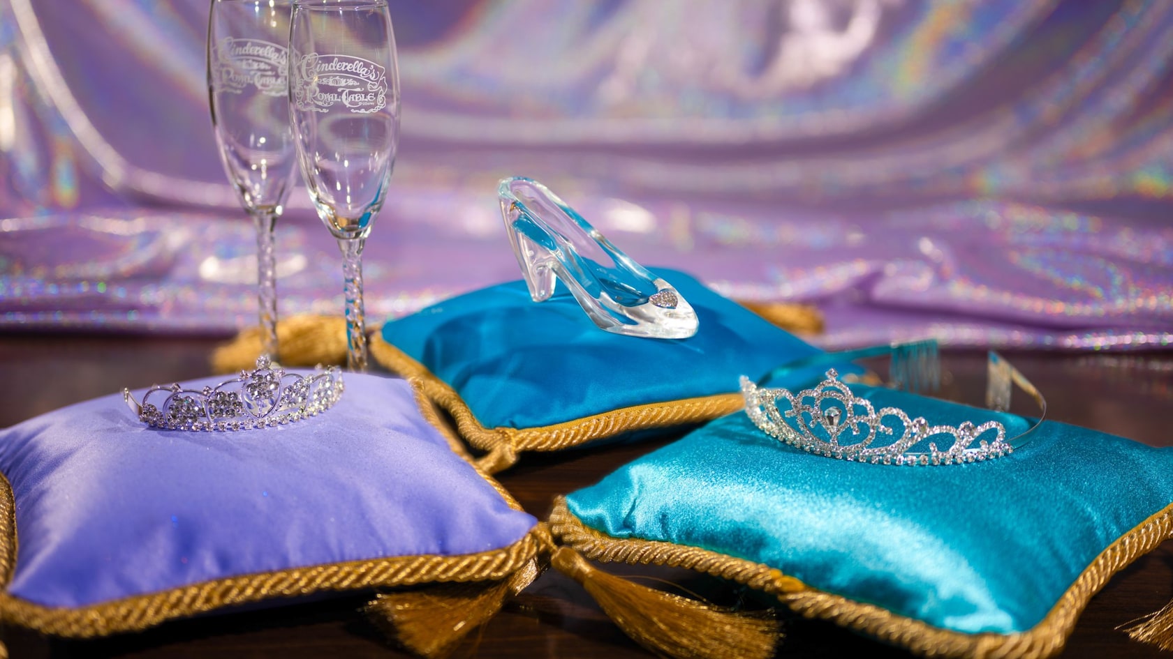 A Cinderella themed tiara, glass slipper and 2 champagne flutes each rest on 3 luxurious pillows