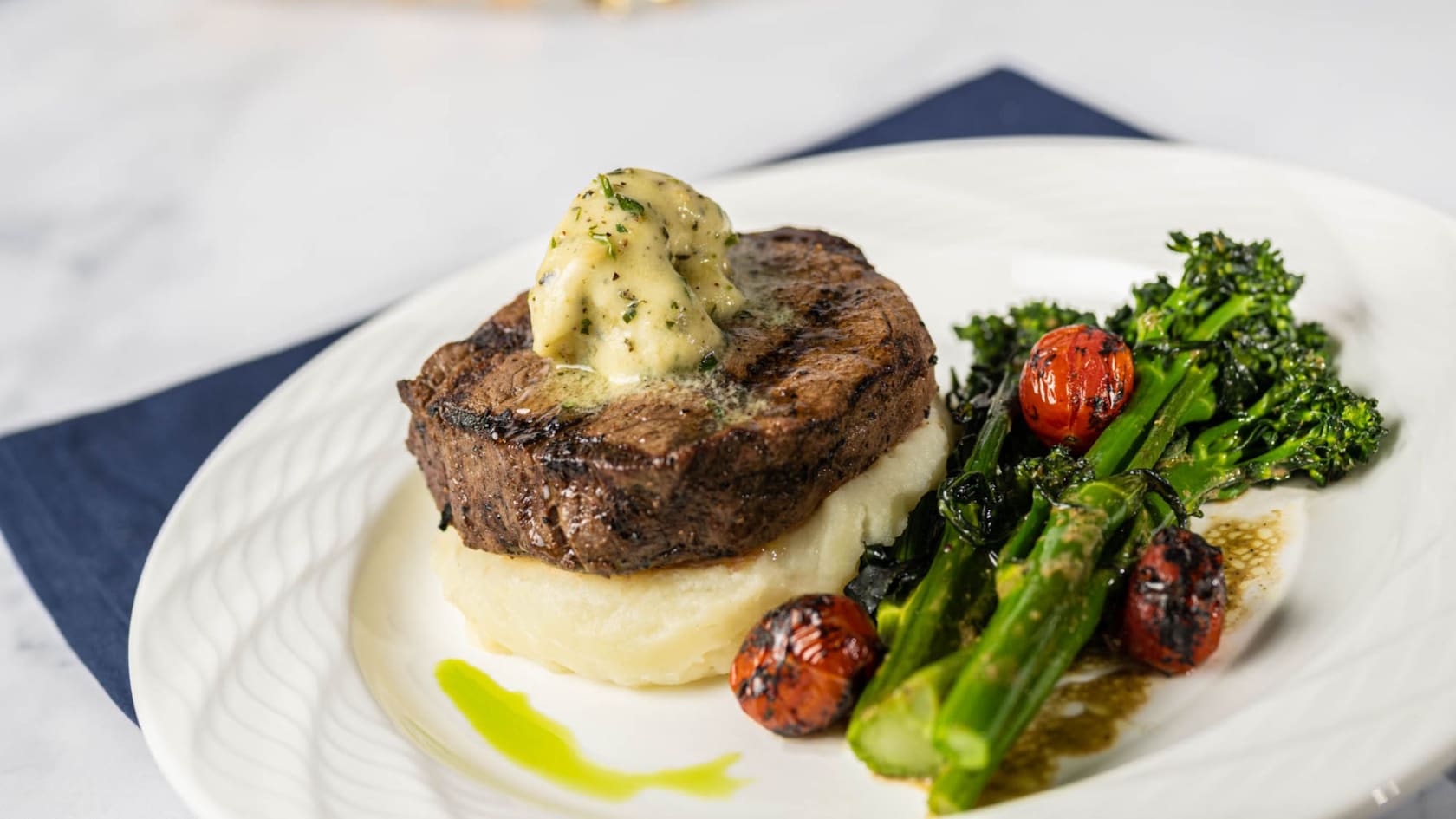 A plated Grilled Filet Mignon featuring a grilled steak topped with butter, served on mashed potatoes with roasted broccolini and cherry tomatoes