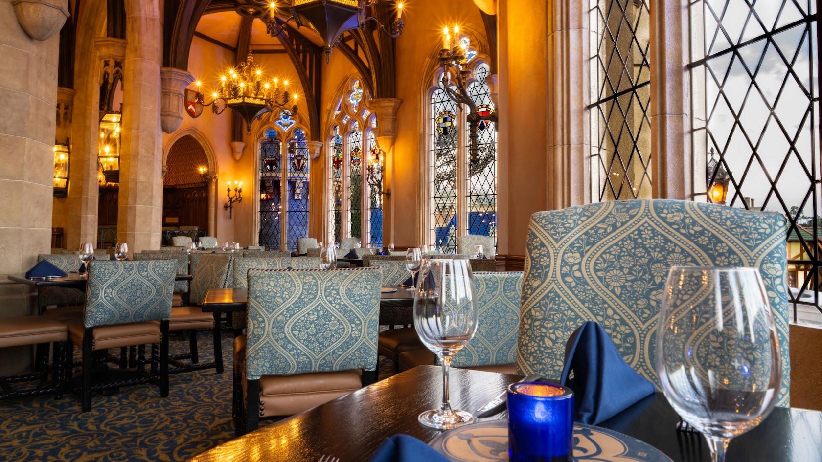 A dining room with arched ceilings, chandeliers, stained glass windows and tables set with napkins and glassware