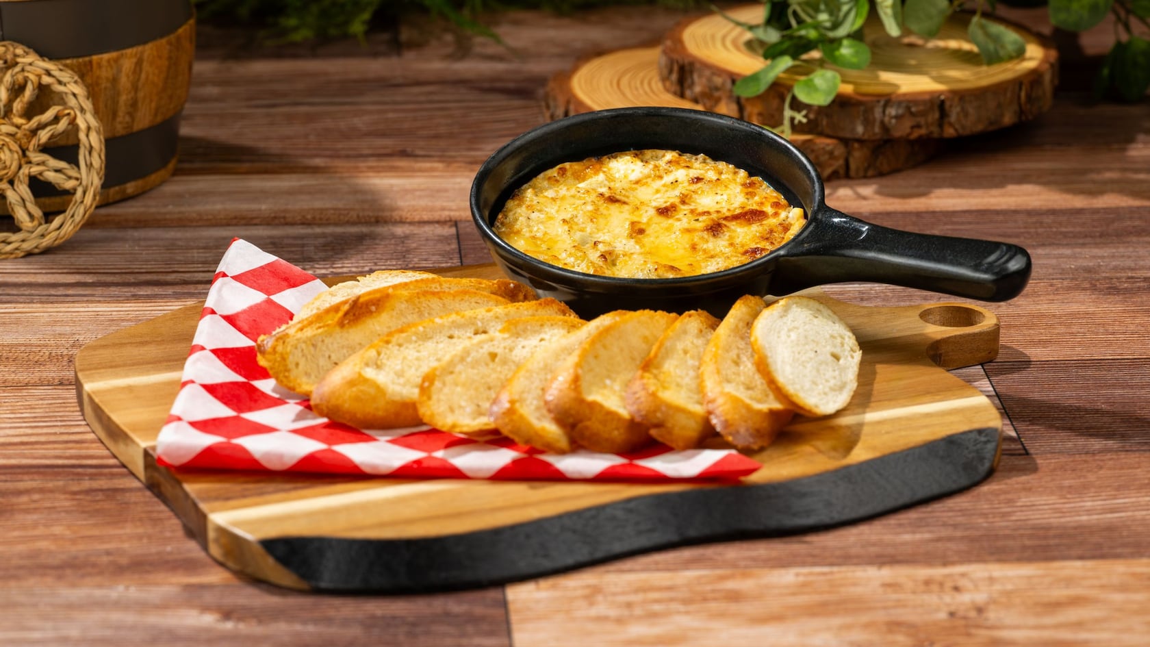 A cast iron skillet of artichoke and crab dip served on a wooden board with slices of toasted baguette