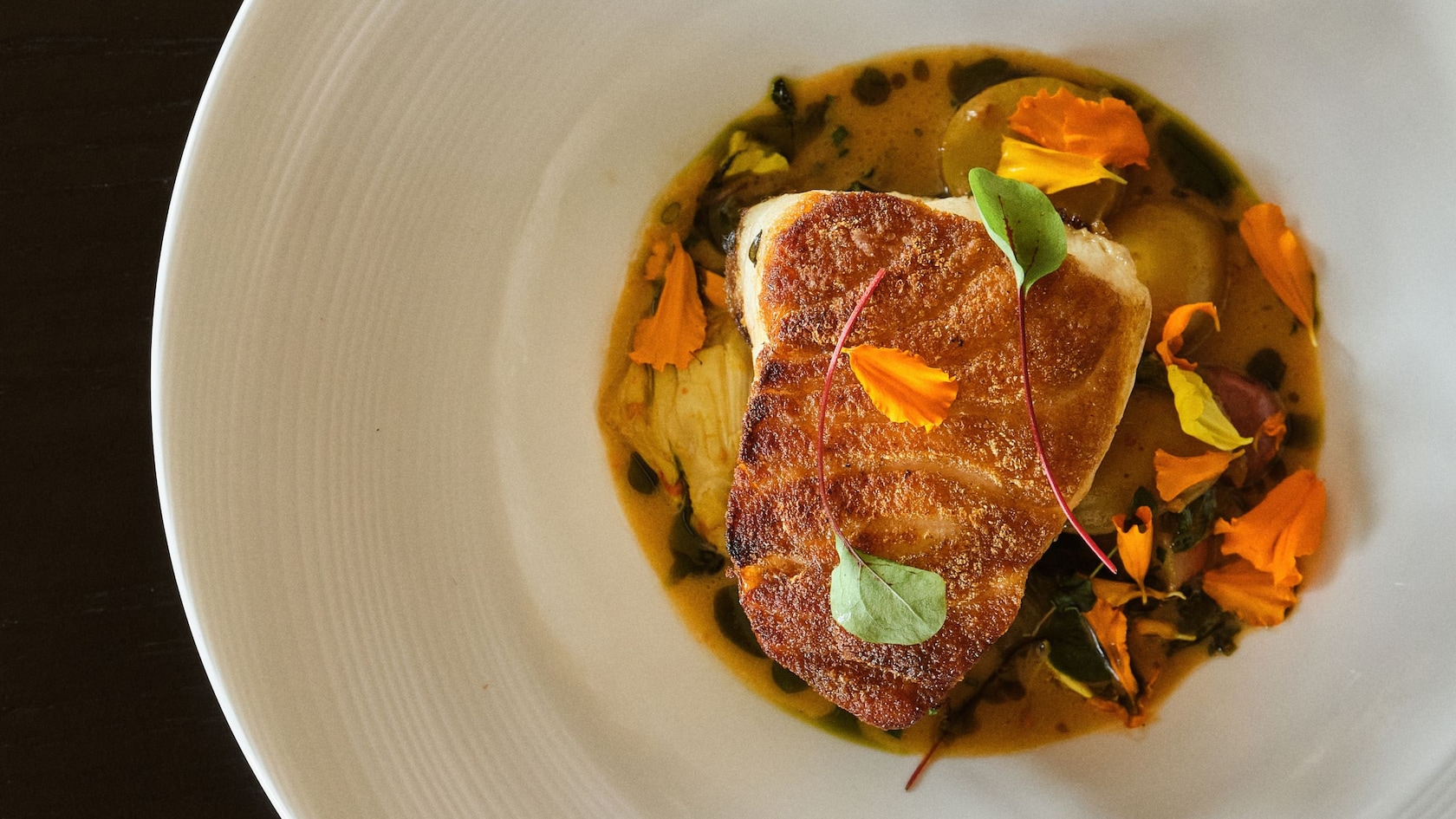A seared fish fillet, surrounded by sauce and garnished with edible flowers and microgreens