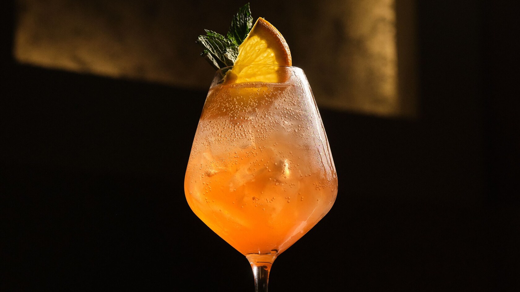 An iced cocktail in a stemmed wine glass garnished with an orange wedge and mint leaves